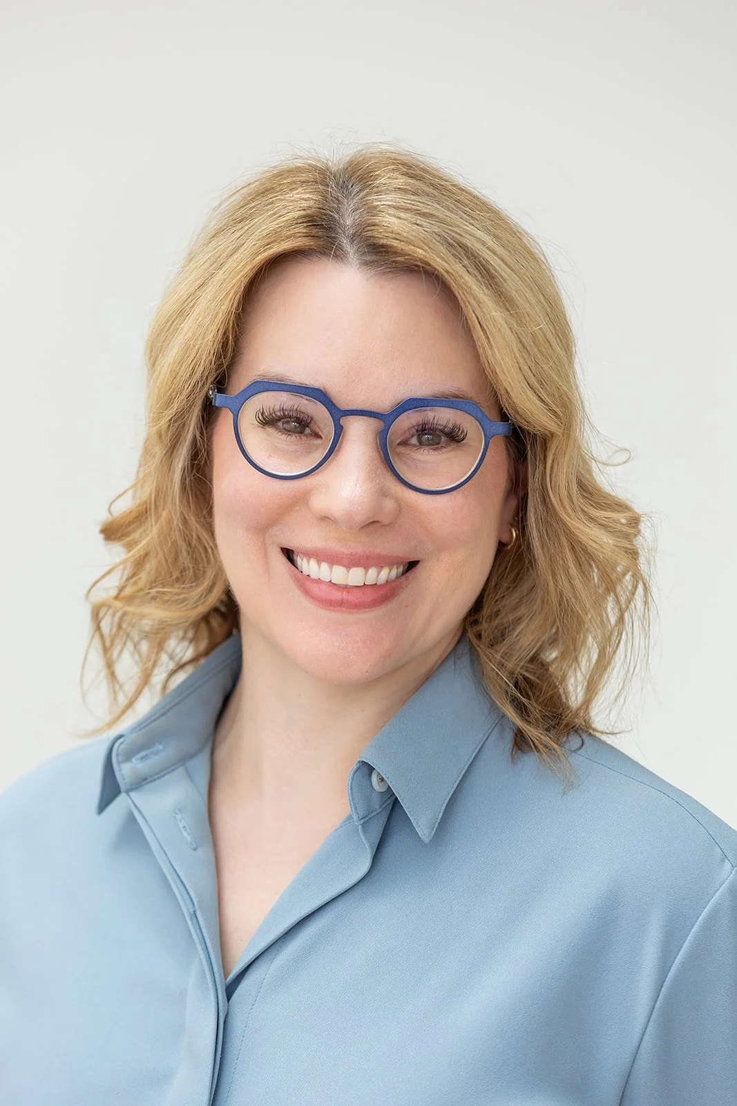 A woman with blonde wavy hair wearing blue glasses and a light blue button-up shirt, smiling at the camera against a plain background.