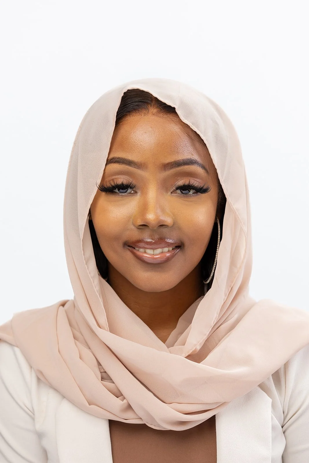 A smiling woman wearing a beige headscarf and earrings against a white background.
