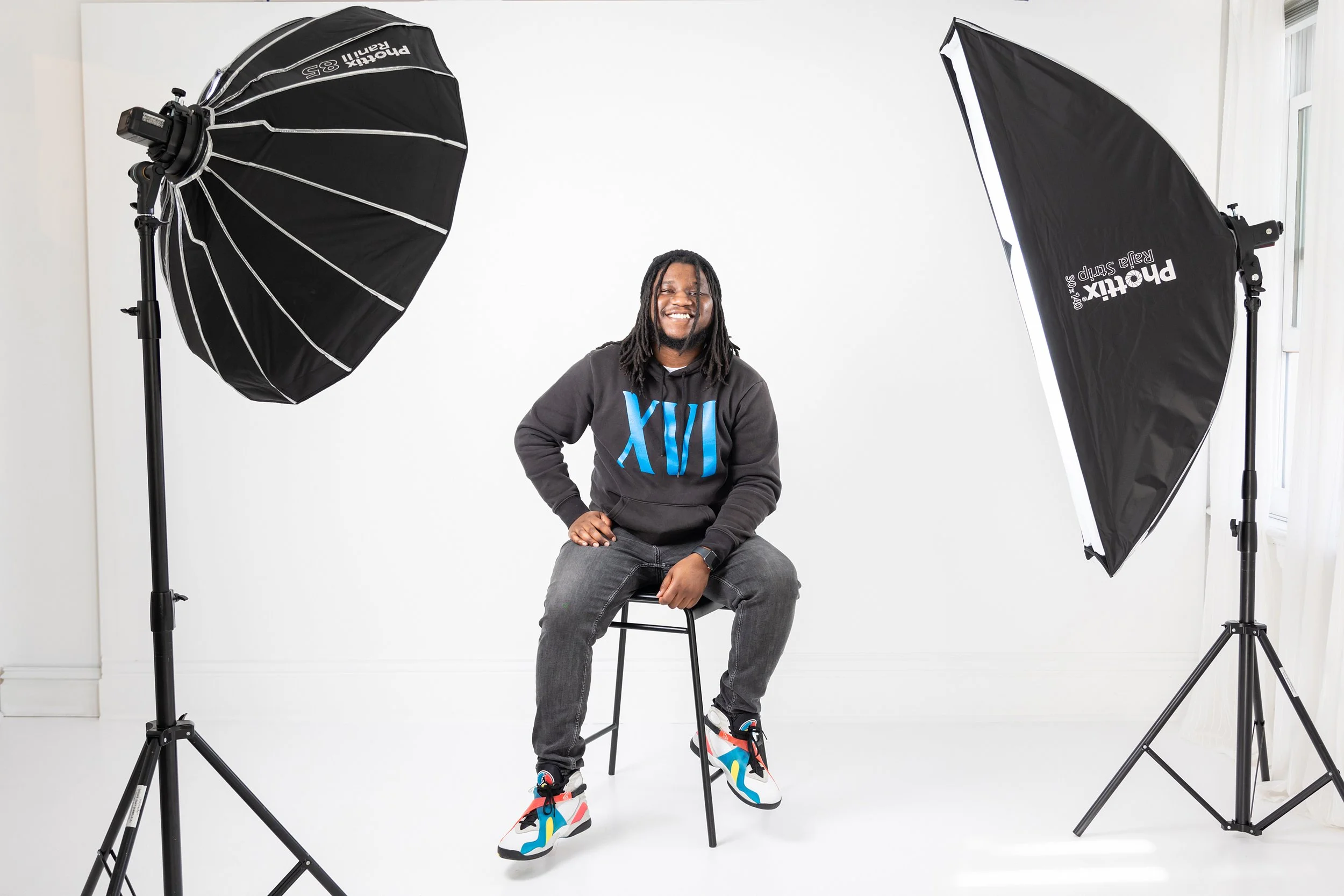A man sitting on a stool in a photography studio with two softbox lights on either side, smiling at the camera. He is wearing a black hoodie with Roman numerals XIII in blue, gray jeans, and colorful sneakers. The studio has a white background and na
