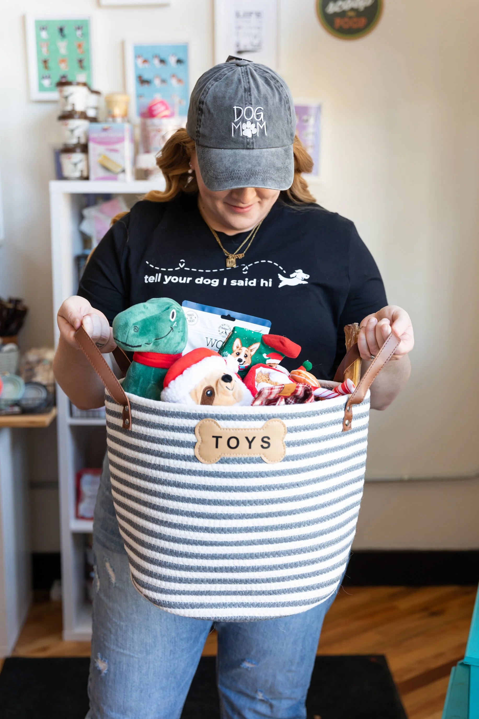 A woman wearing a gray cap and black t-shirt holding a striped storage basket filled with plush toys and dog-themed items, inside a store with shelves of pet supplies.