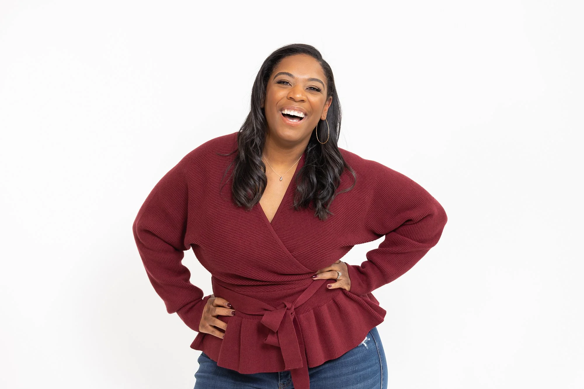 A woman with dark hair smiling, wearing a red wrap sweater and jeans, standing against a white background.