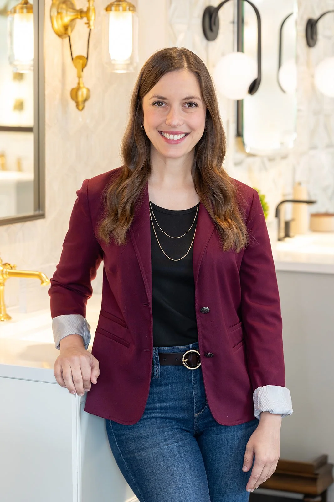 A woman with long brown hair, wearing a maroon blazer, black shirt, and blue jeans, smiling while standing in a well-lit bathroom or kitchen with gold fixtures and white countertops.
