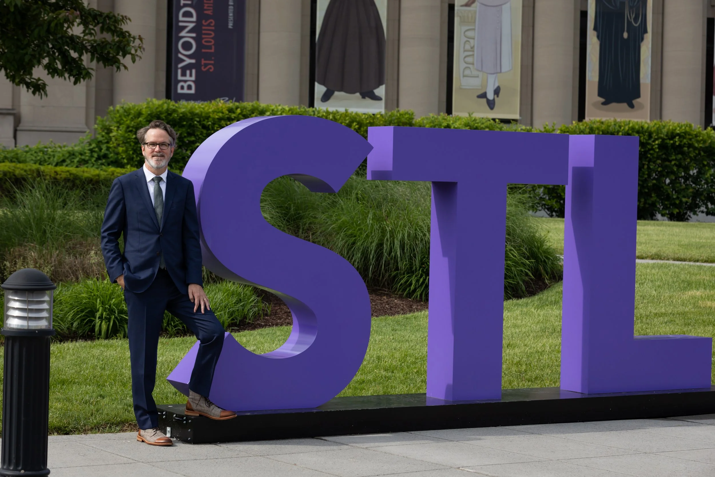 A man in a navy suit and glasses standing outdoors next to a large purple sign spelling 'ST' with a building and banners in the background.
