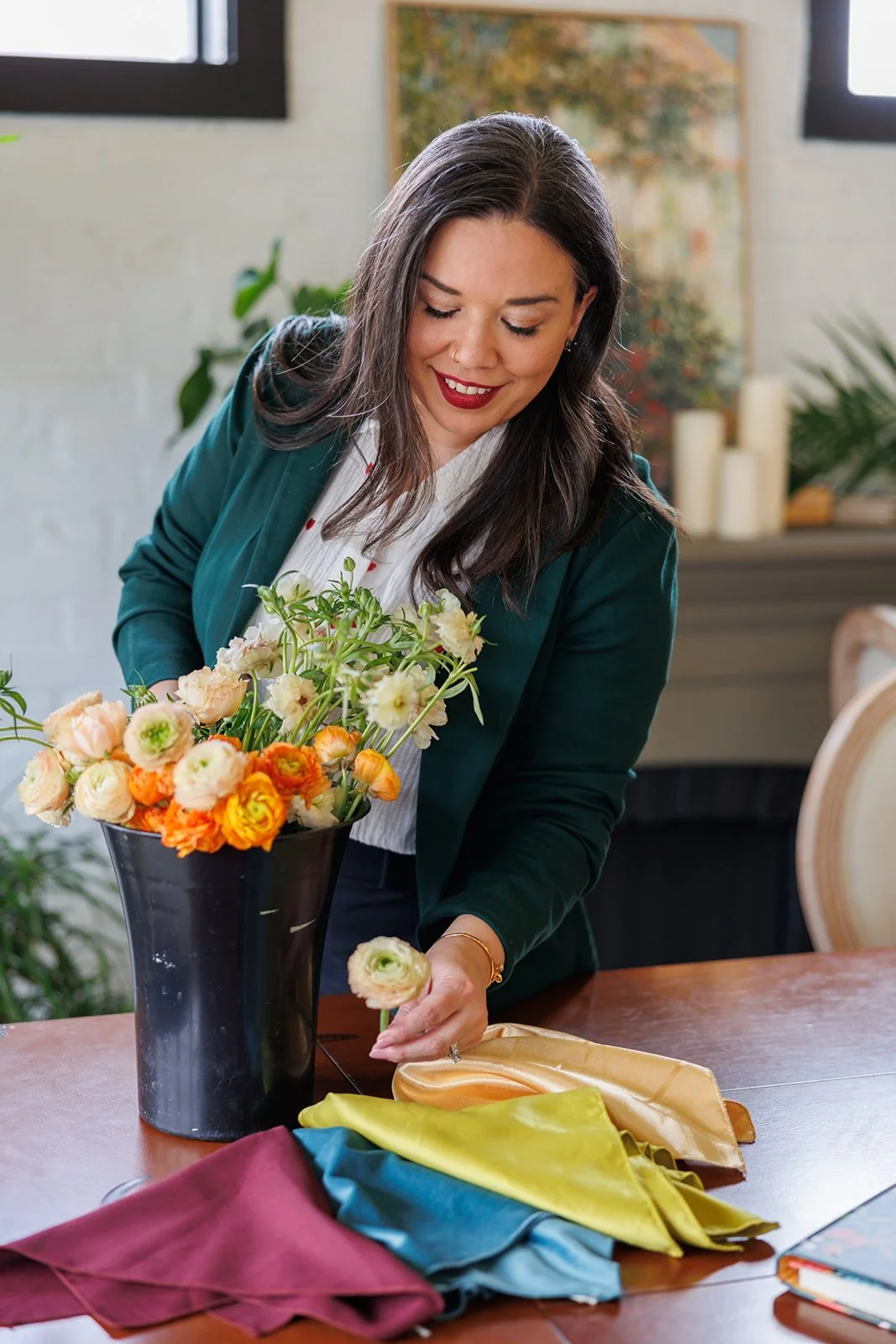A woman arranging a bouquet of flowers in a black vase on a wooden table, with colorful fabric swatches nearby, in a well-lit room with plants and candles in the background.