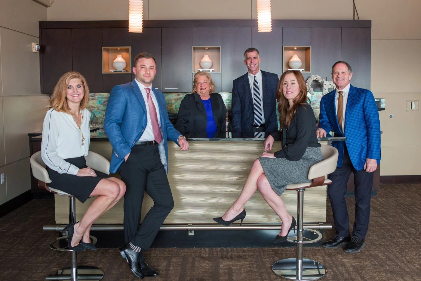 Group of six professionals in business attire posing in modern office kitchen with bar counter.
