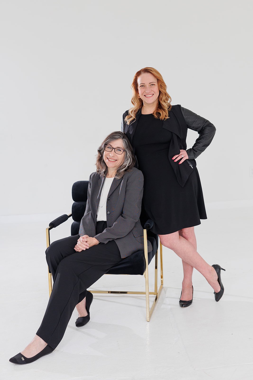 Two women, one sitting in a black chair and the other standing behind her, smiling in a studio with a plain white background.