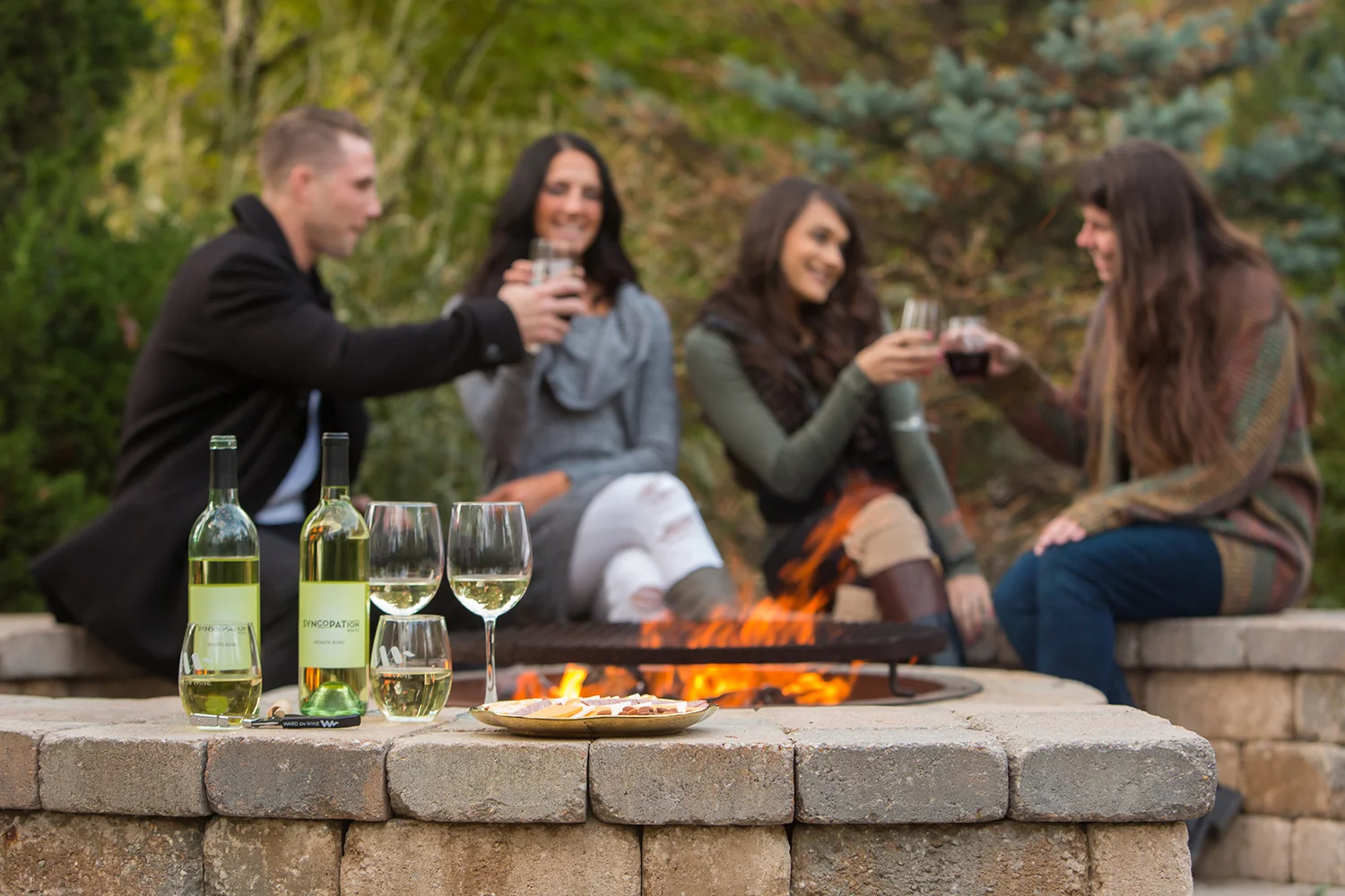 Four people sitting around a fire pit and enjoying drinks outdoors. In the foreground, there are two bottles of white wine, two glasses of white wine, two glasses of red wine, and a plate of cheese on a brick table.