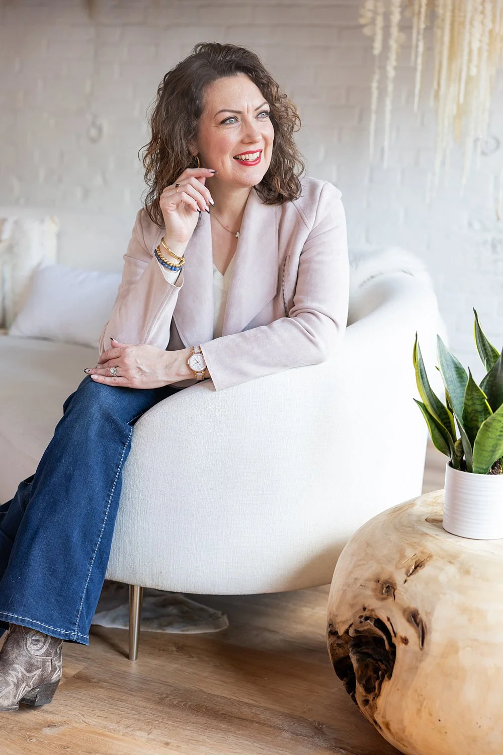 A woman with curly hair sitting on a white sofa, smiling and looking to her left, with a plant on a wooden table beside her, in a bright room with white walls and hanging decor.