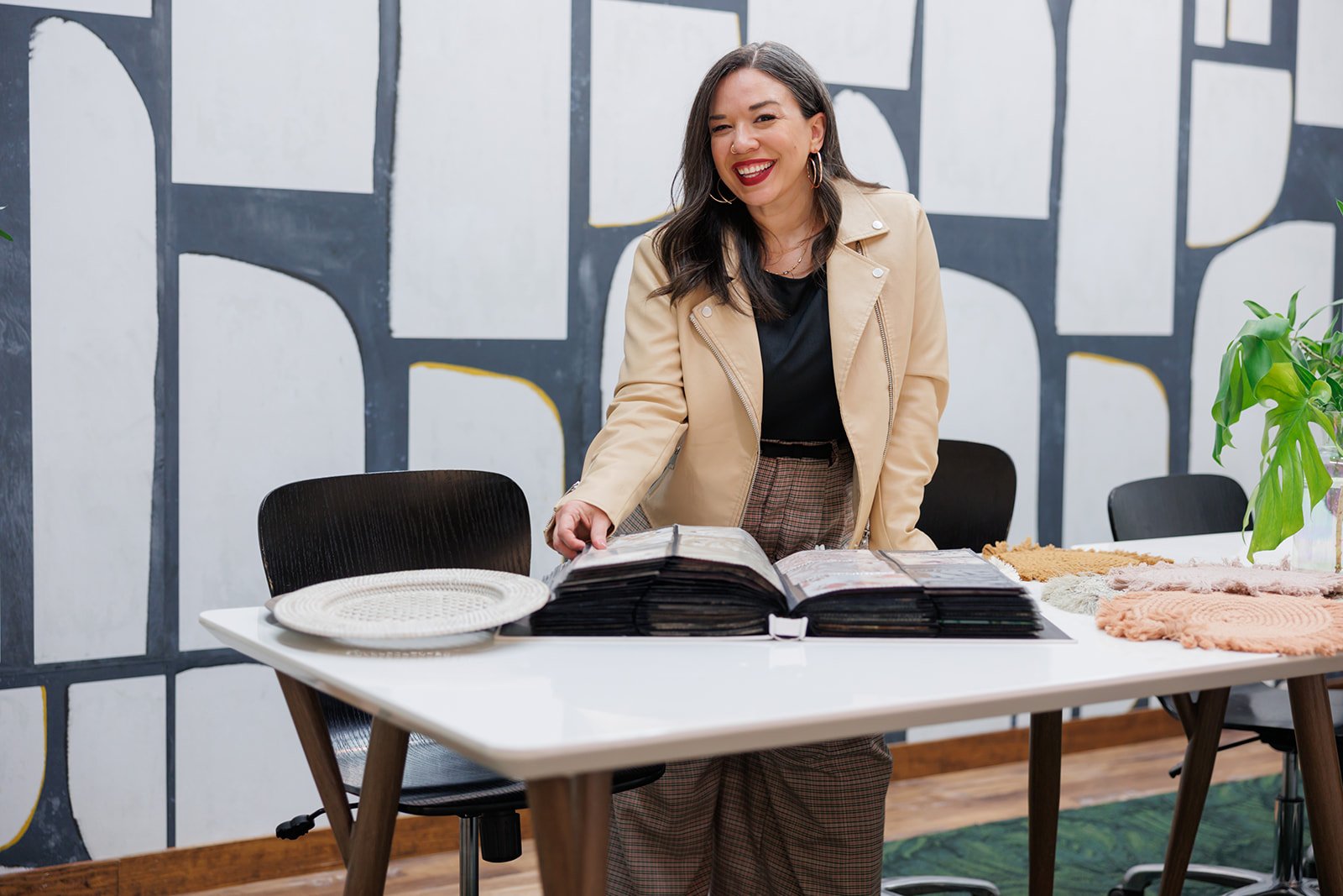 A woman with dark hair, wearing a beige jacket and plaid pants, is smiling and looking at a color sample book on a table, with decorative woven items and a potted plant in the background.