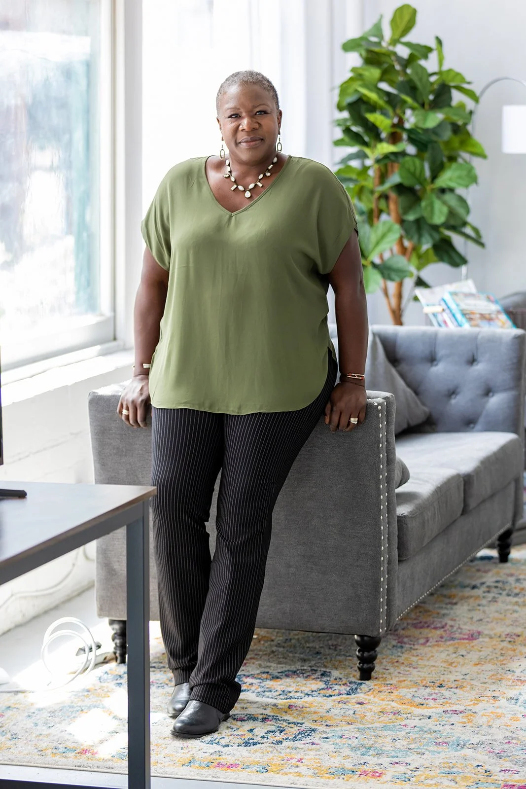 A woman standing in a living room, wearing a green blouse, black pinstripe pants, and black shoes. She is accessorized with a necklace, bracelet, ring, and earrings, and is standing next to a gray sofa.