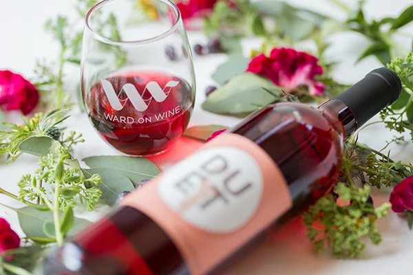 A glass of red wine and a bottle of wine on a table decorated with flowers and green leaves.