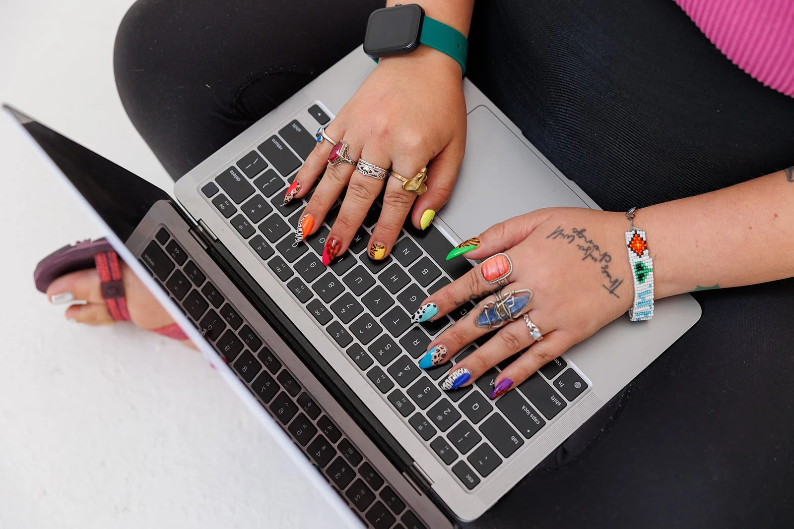 Person using a laptop with colorful painted nails, wearing rings, bracelets, and an Apple Watch, sitting on a white surface.