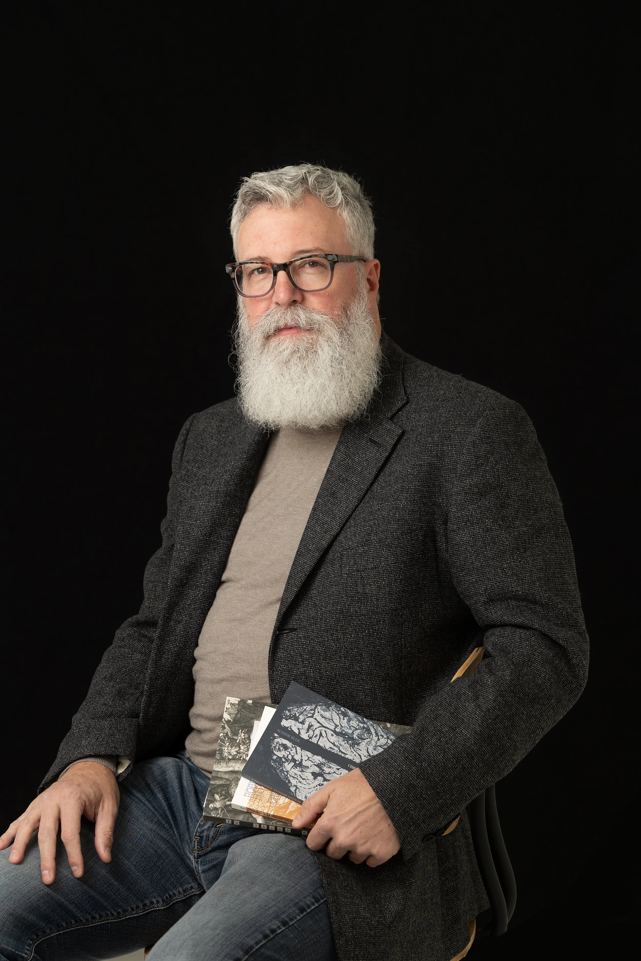 An older man with a white beard and glasses wearing a dark blazer and gray t-shirt, sitting against a black background, holding books in his lap.