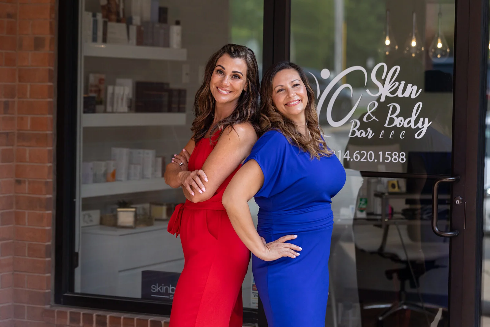 Two smiling women standing outside a skin and body care business, one in a red dress and the other in a blue dress, with the business's glass door and signage in the background.