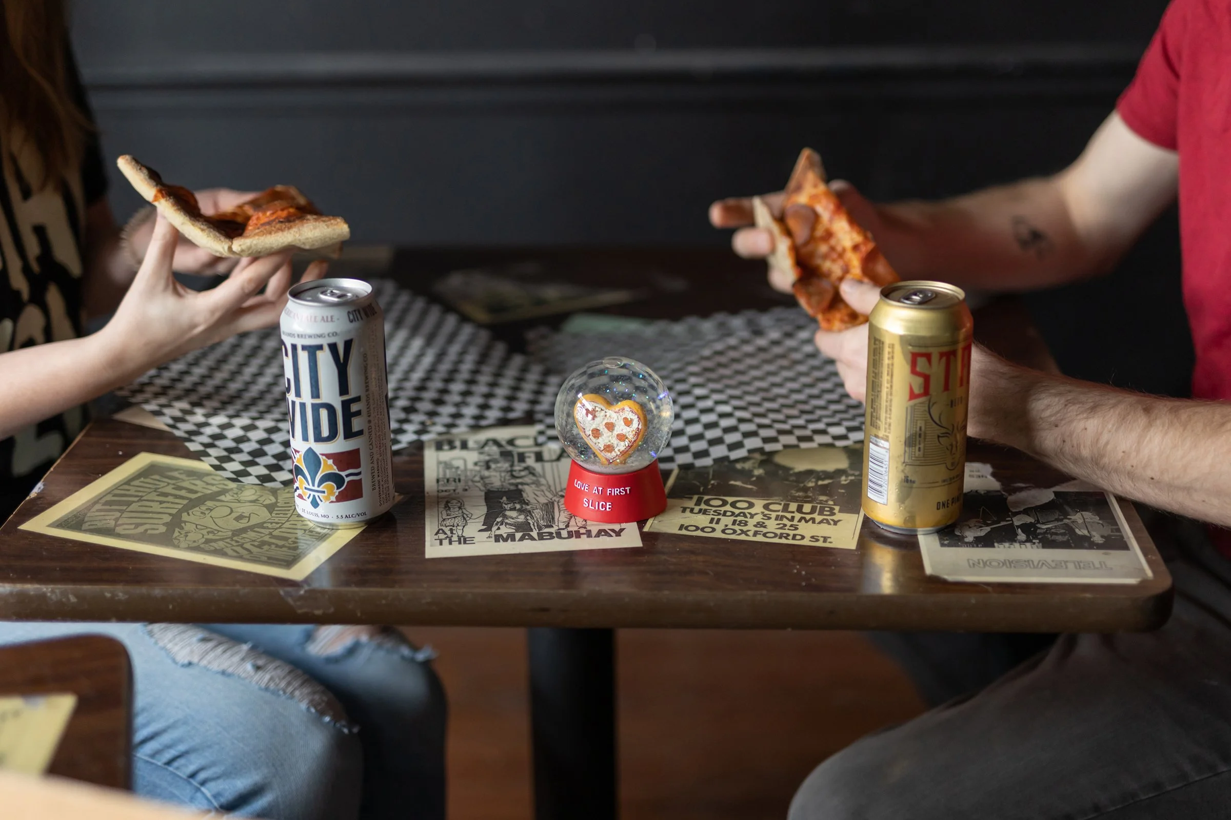 Two people sitting at a wooden table, eating pizza and drinking canned beverages, with posters and a snow globe with a heart-shaped cookie inside on the table.