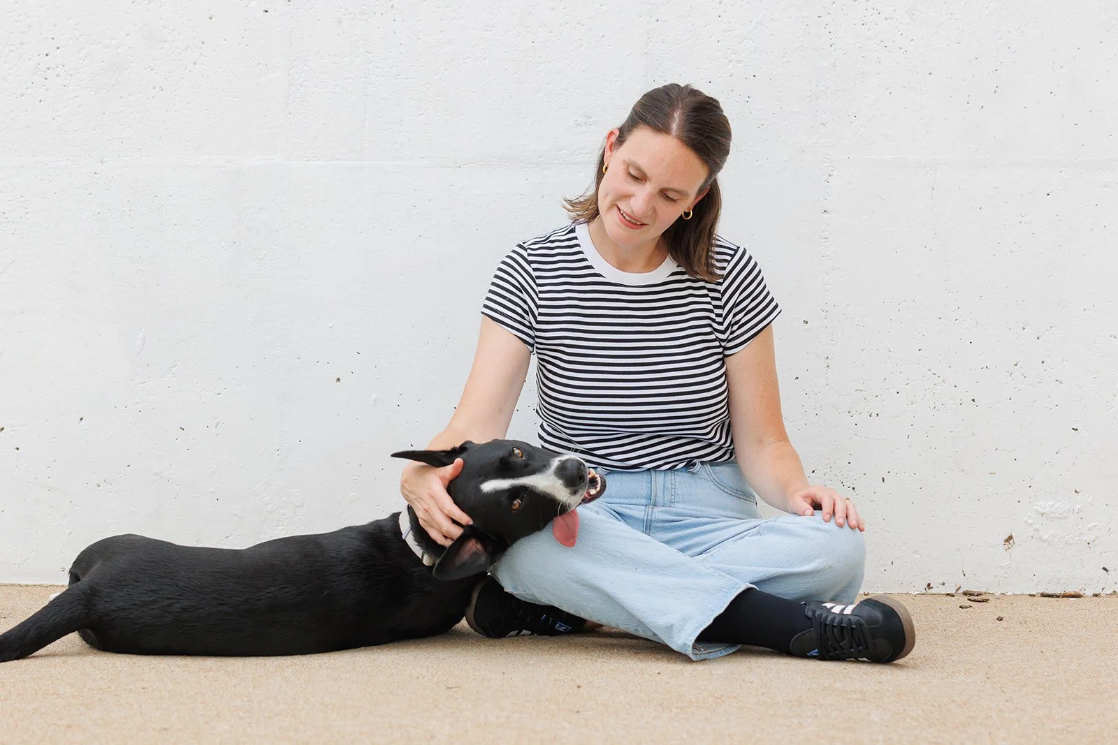 Young woman sitting on the floor, petting a black and white dog with her right hand, against a white textured wall.