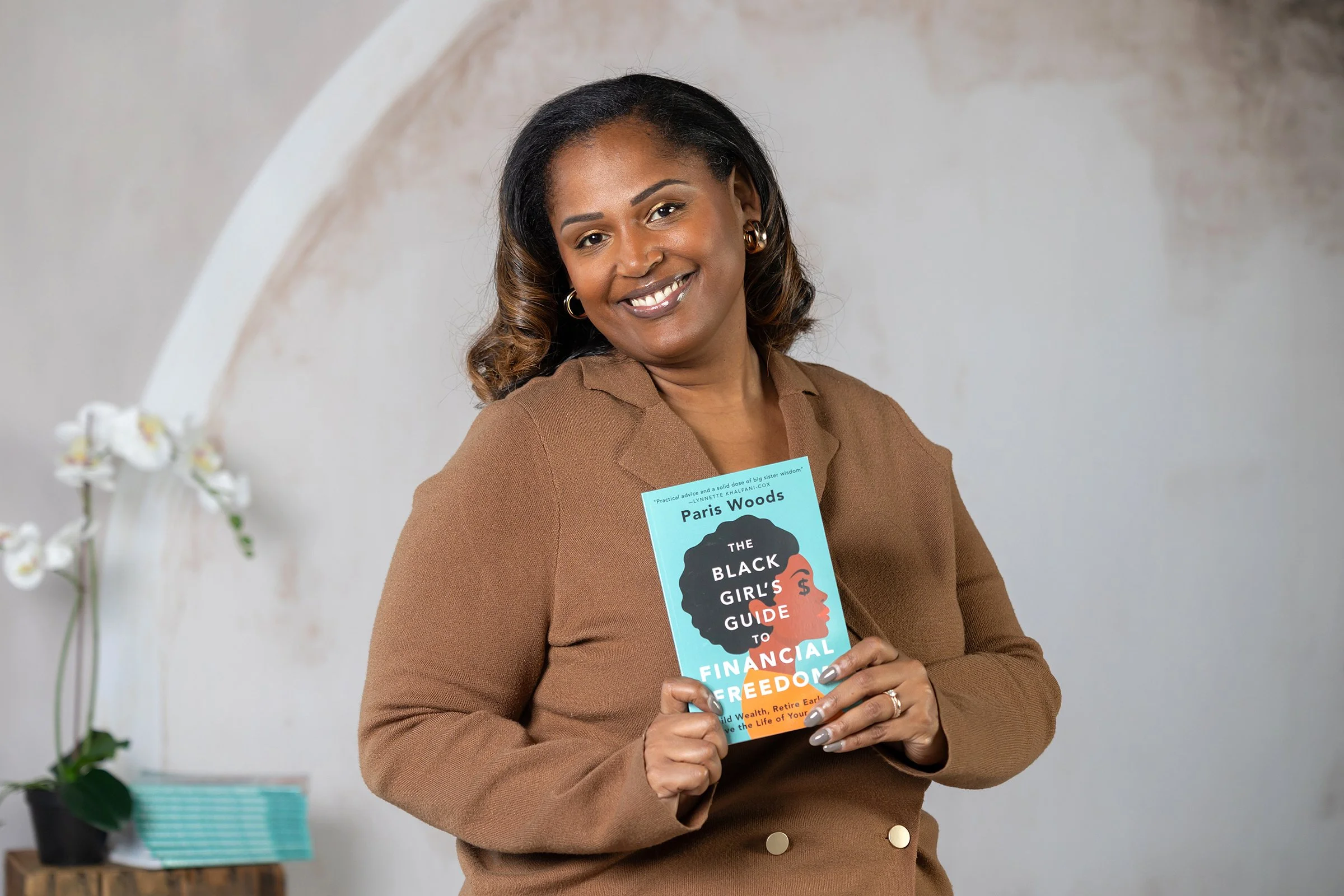 A woman with medium brown skin, curly black and brown hair, and gold hoop earrings, smiling and holding a book titled 'The Black Girl's Guide to Financial Freedom' in front of a plain light-colored wall, with a potted orchid and a stack of books on a small table in the background.