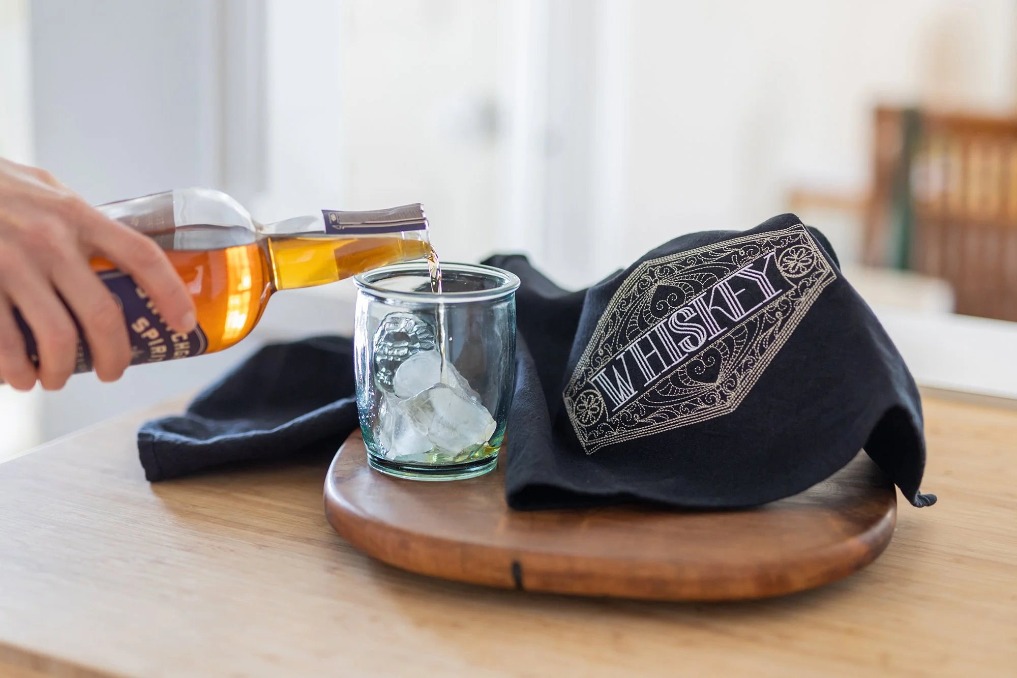 A person pouring whiskey into a glass with ice on a wooden table, with a black cloth and a black bandana with white embroidery reading 'WISHWIT' nearby.