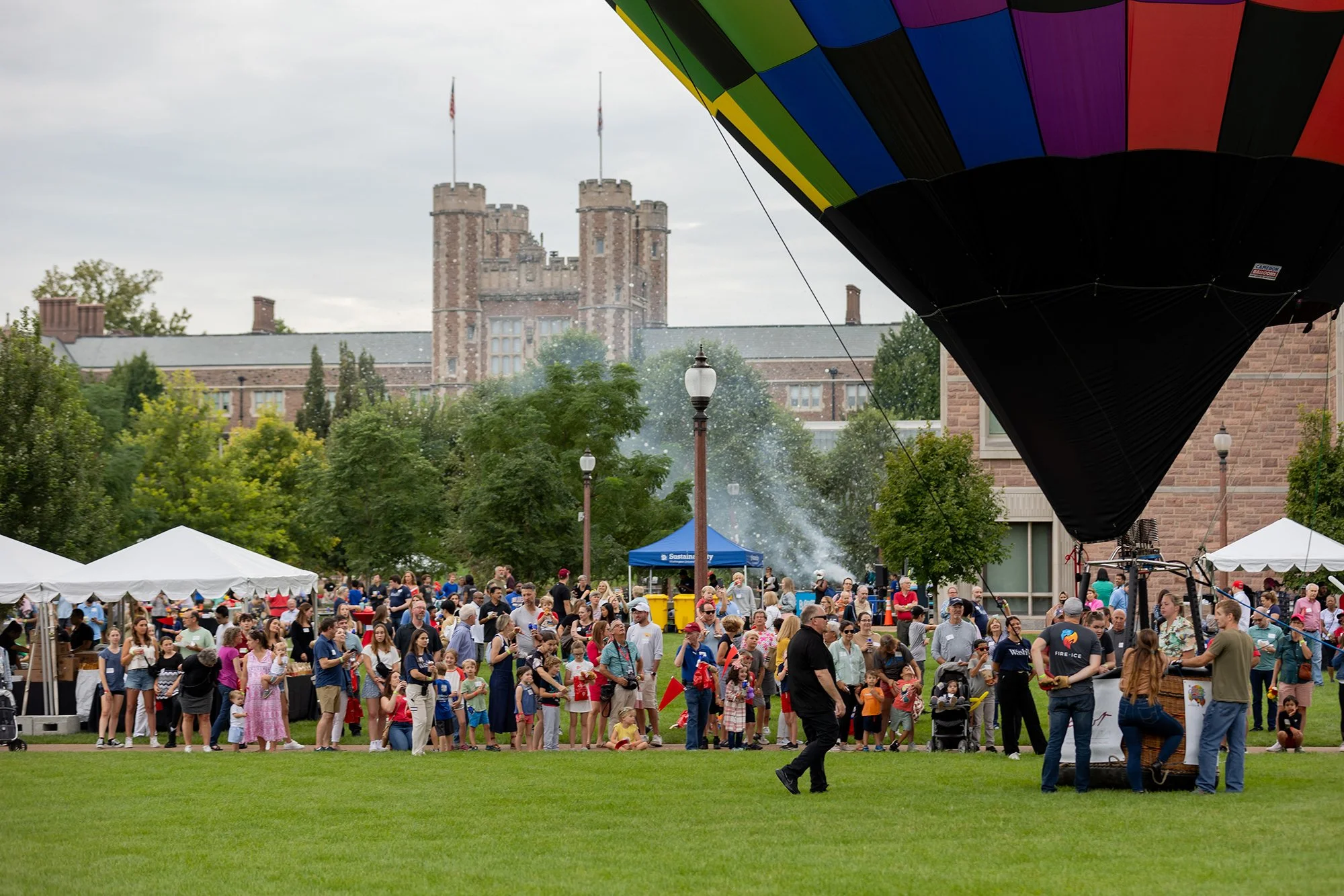Crowd gathered at an outdoor event with tents, a hot air balloon, and a castle in the background on a cloudy day.