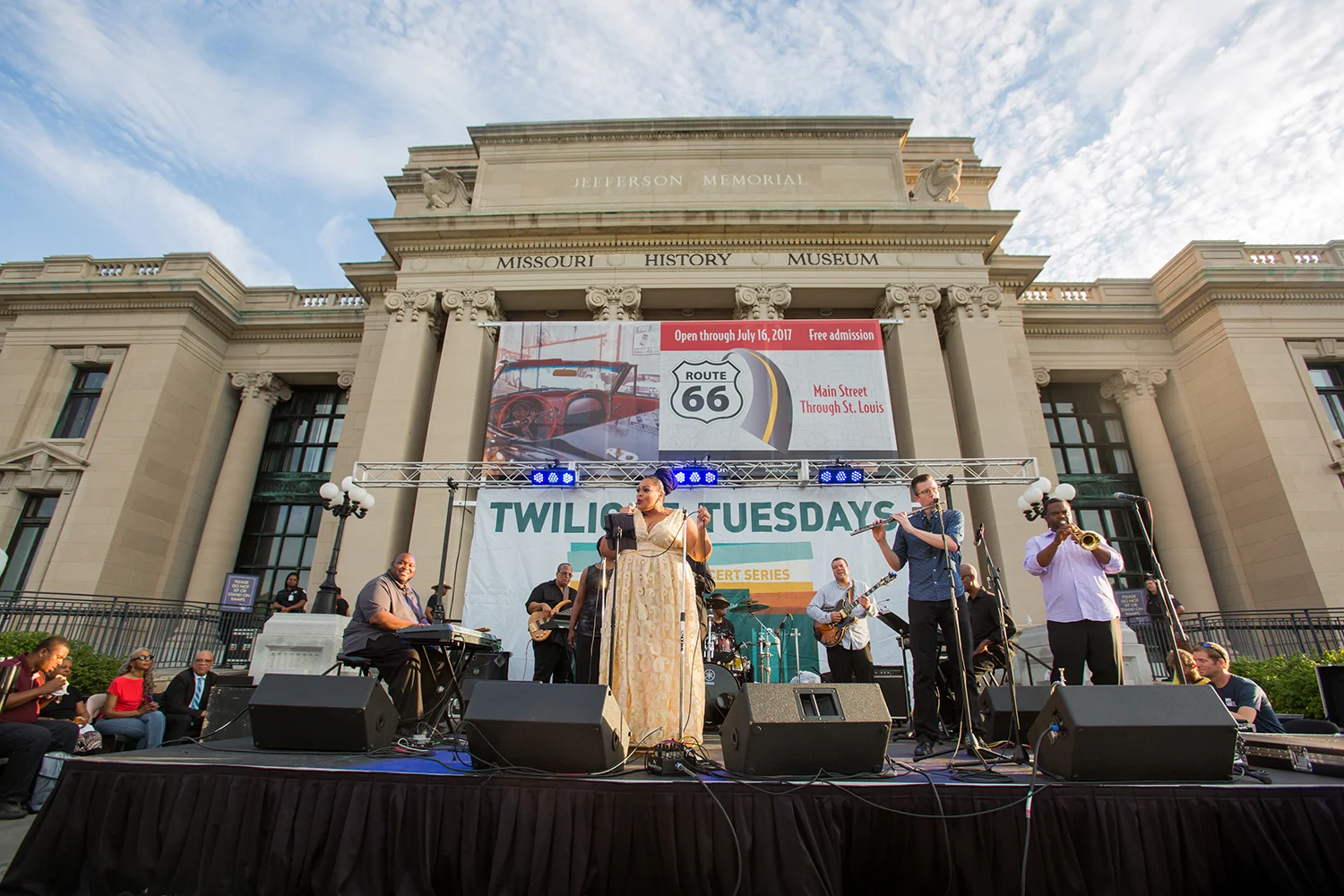 A live music performance on a stage in front of the Missouri History Museum; band members including a female singer, keyboardist, and horn players, with an audience seated around.