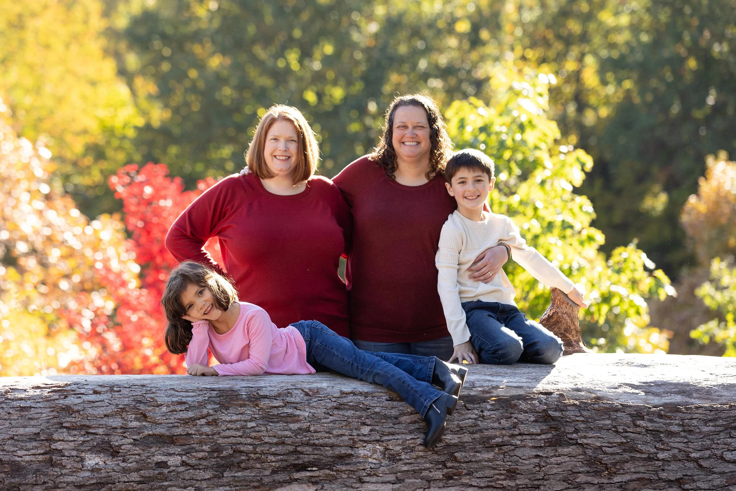 Family of four with two women and two children, sitting on a large fallen tree trunk outdoors during fall, with colorful autumn leaves in the background, laughing and smiling.
