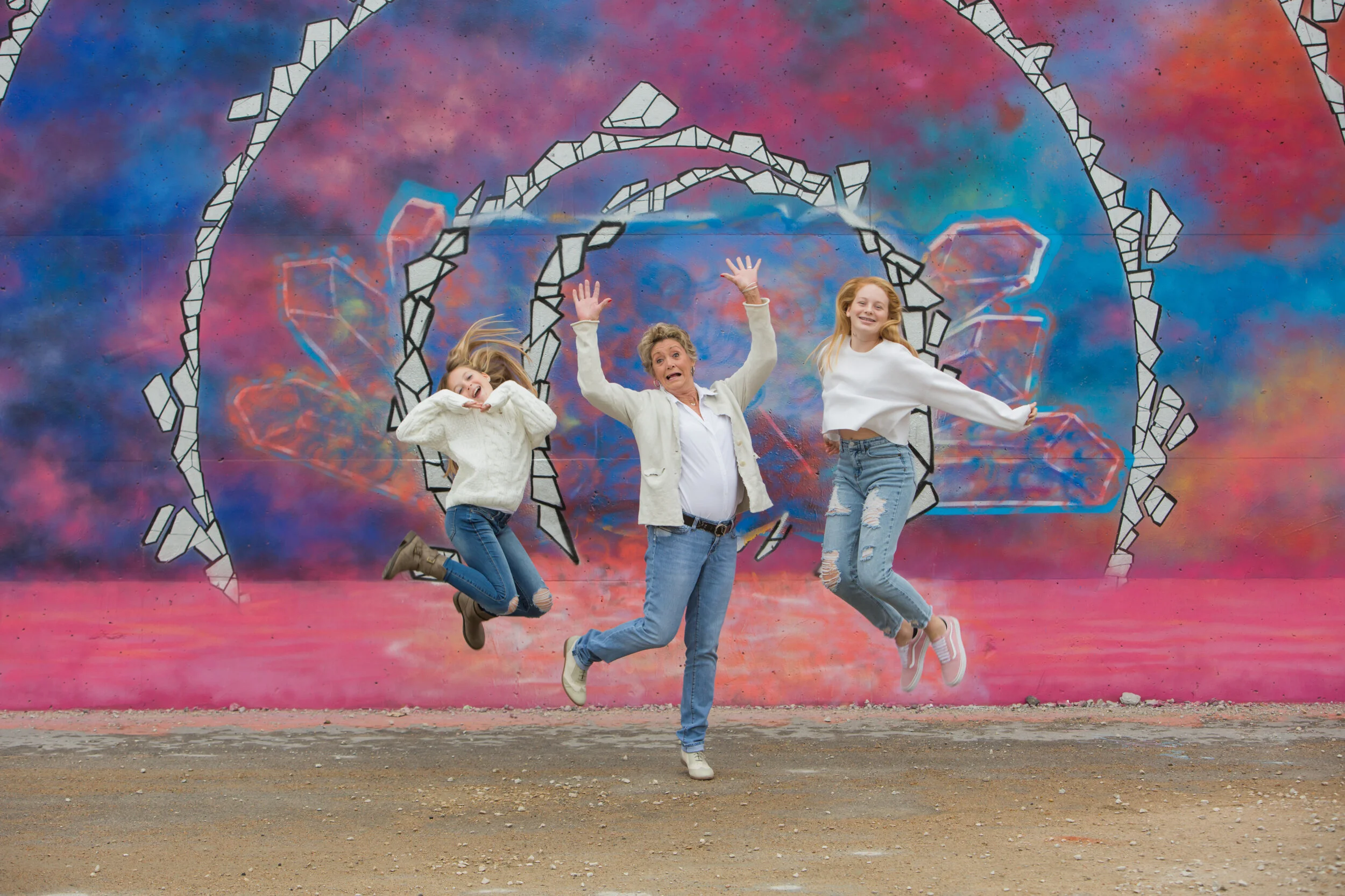Three people jumping in front of a colorful graffiti mural featuring a cosmic background with galaxy-like swirls and geometric shapes.