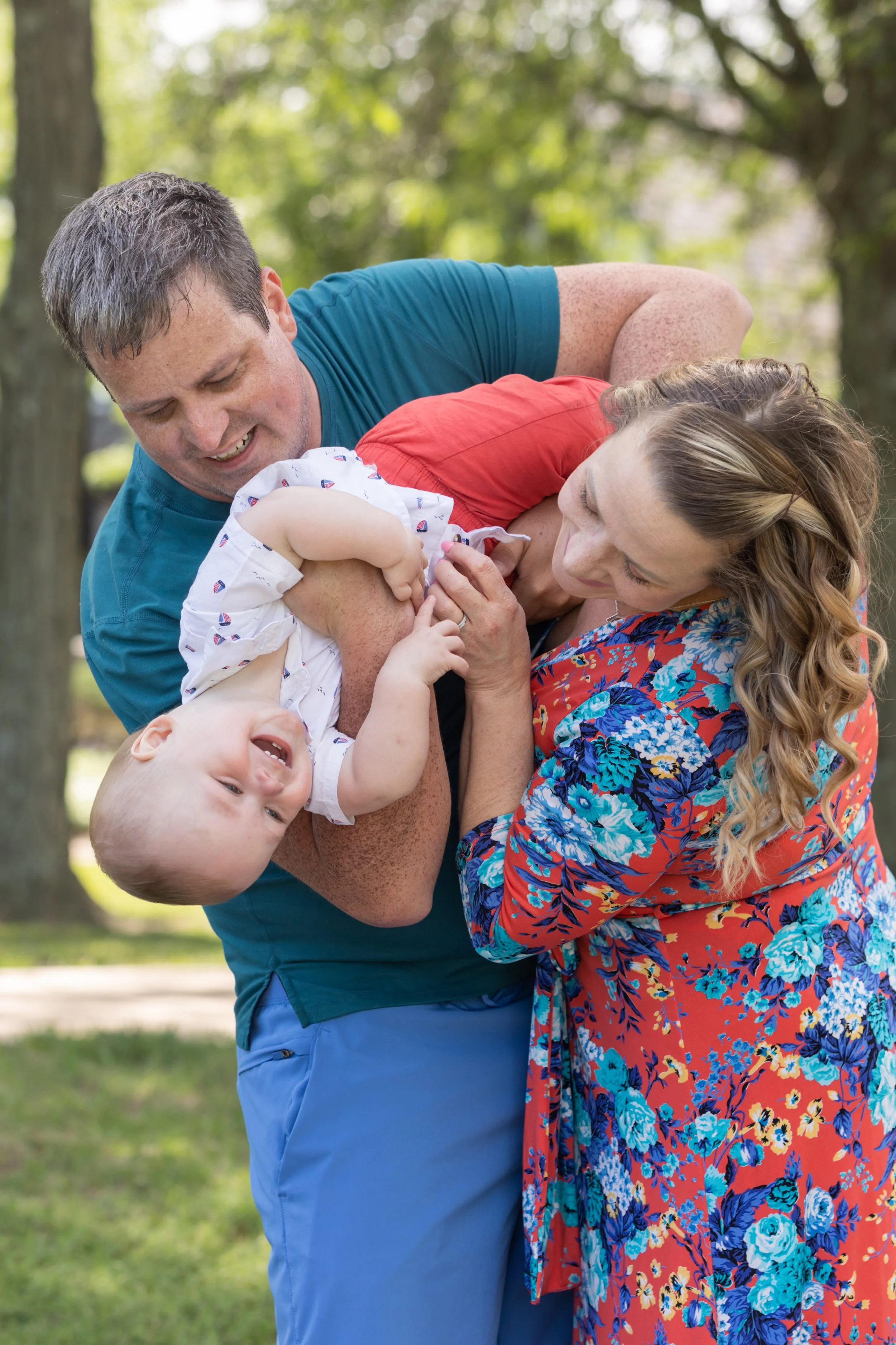 Family outdoors, father, mother, and young child playfully wrestling on grass with trees in the background.