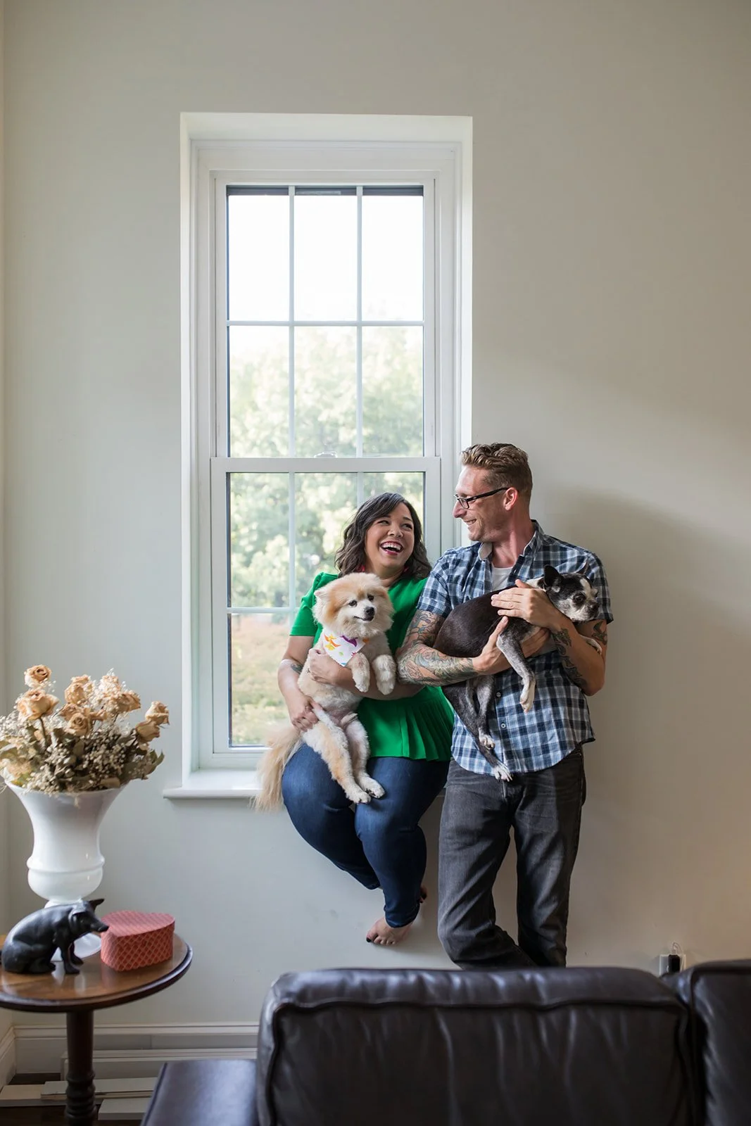 A woman and a man are inside a room near a window, smiling and holding small dogs. The woman is wearing a green top and jeans, holding a fluffy white dog. The man is wearing glasses, a checkered shirt, and dark pants, holding a black and gray dog. Th