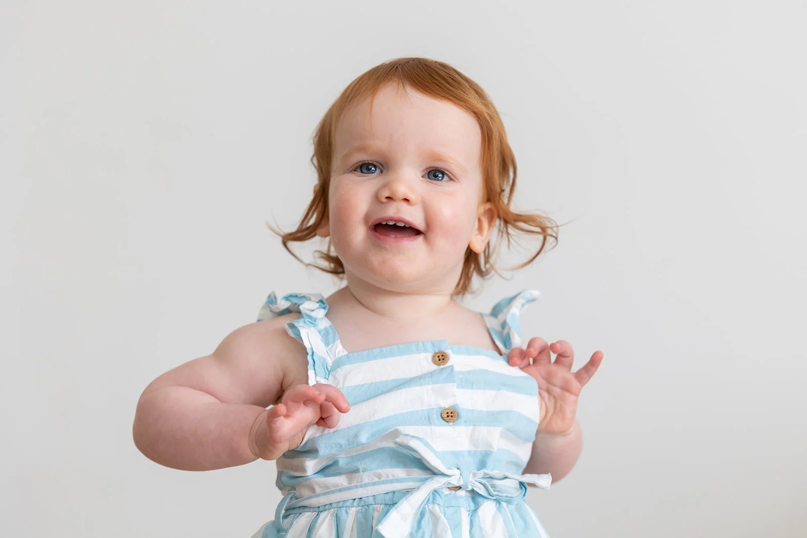 A young girl with red hair and blue eyes wearing a blue and white striped dress with shoulder straps and buttons, standing against a plain background.