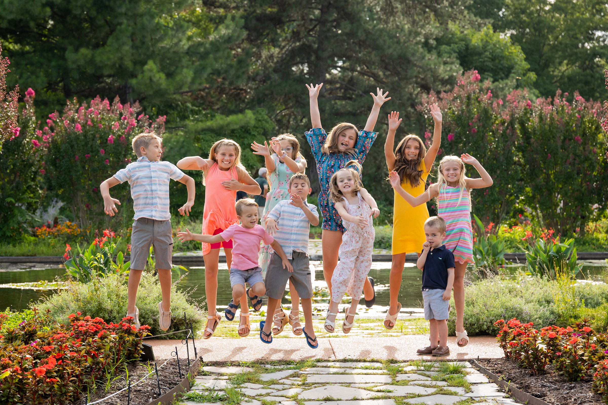 Group of children jumping and playing outdoors in a garden near a pond, surrounded by colorful flowers and green trees.