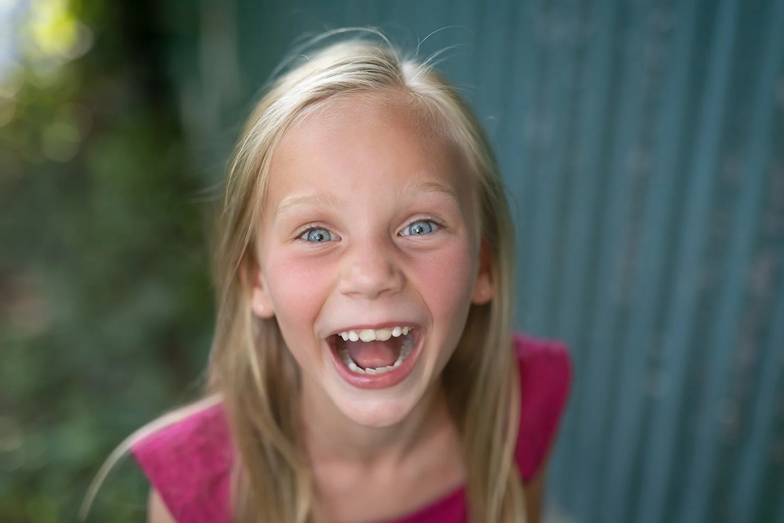 A young girl with blonde hair and blue eyes smiling widely with her mouth open, outdoors near a blue fence and greenery.