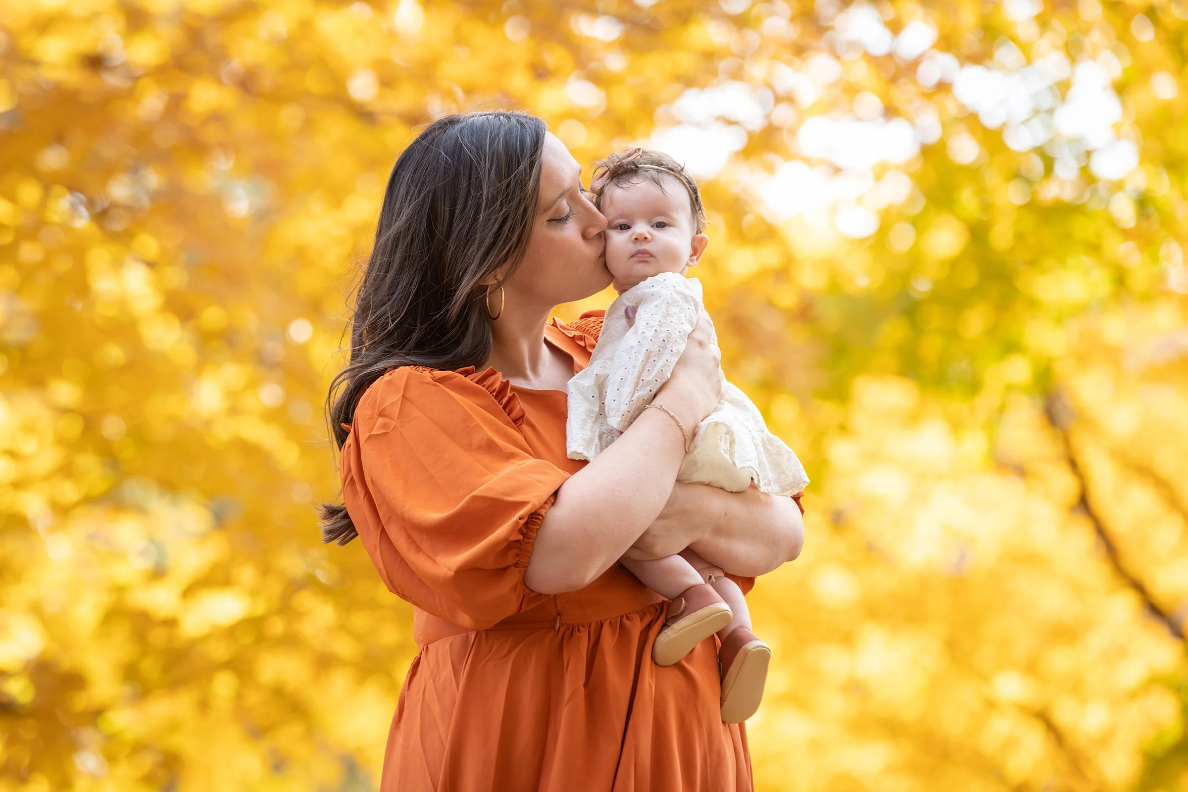 A woman in an orange dress holding and kissing a baby girl in a white dress, with a fall background of yellow and orange leaves.