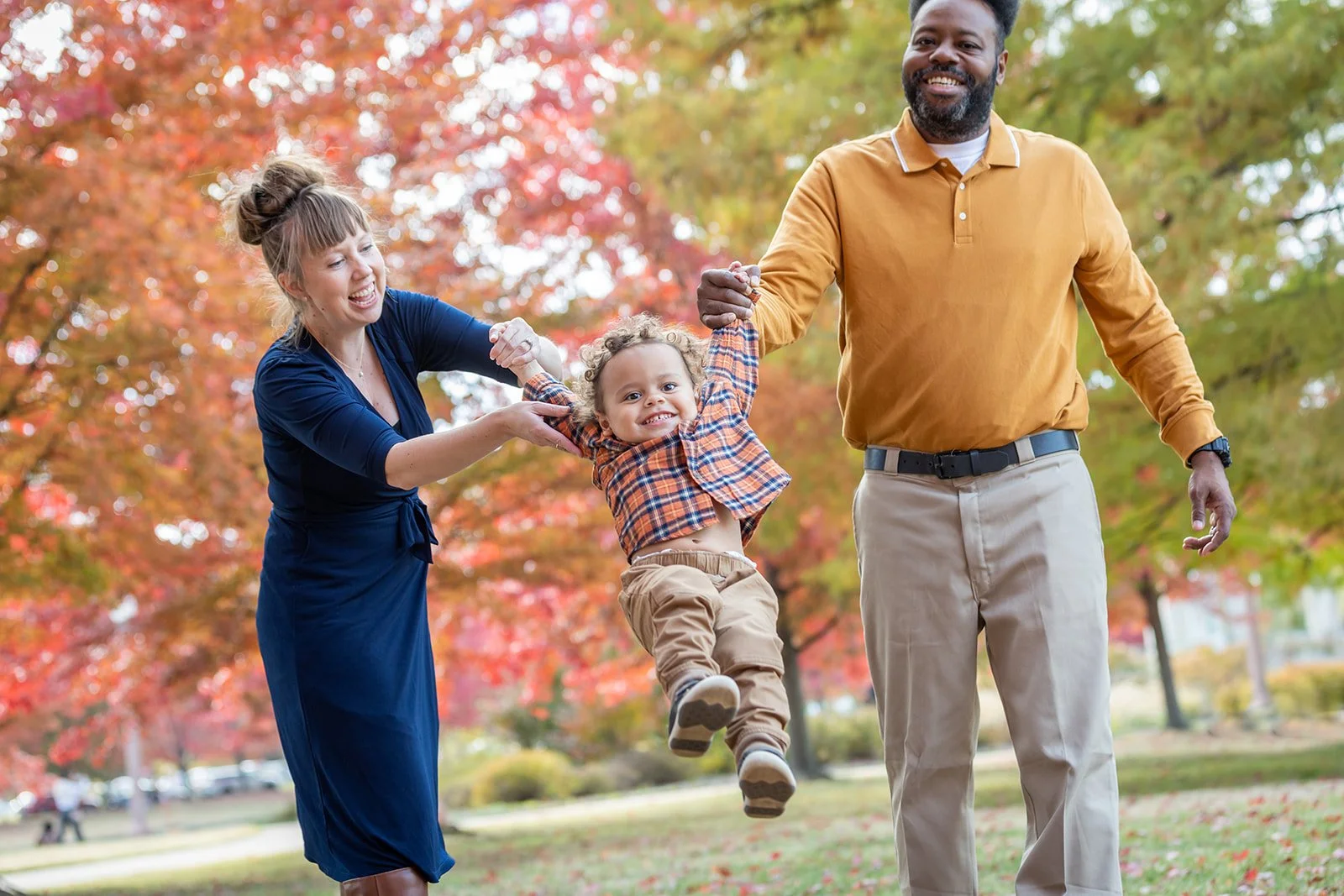 A family of three enjoying a walk in a park during fall, with colorful autumn trees in the background. The mother and father are holding their young child's hands as he swings between them, all smiling and happy.
