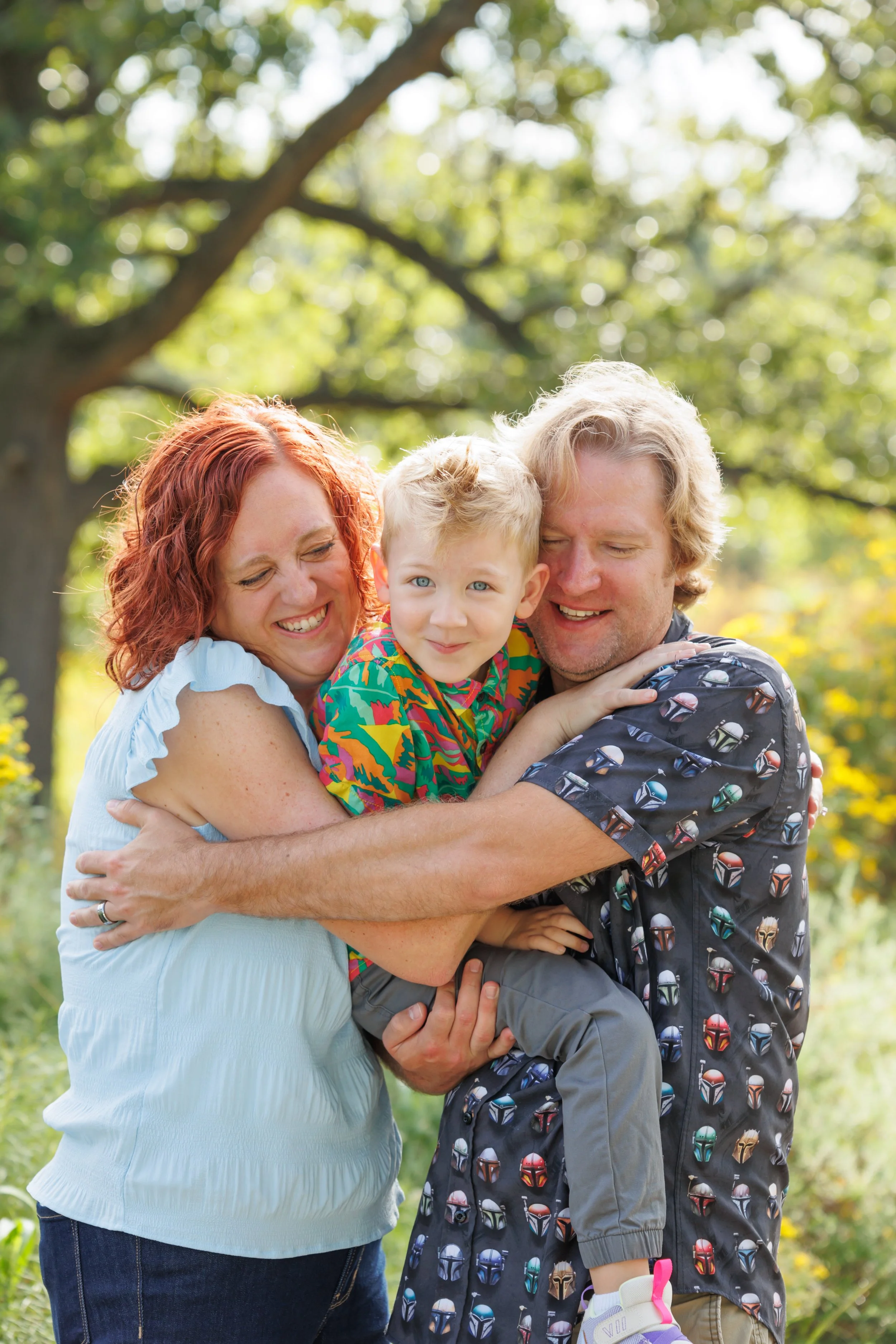 A smiling family of three, a woman with red hair, a man with blond hair, and a young girl with blond hair, hugging outdoors on a sunny day near trees.