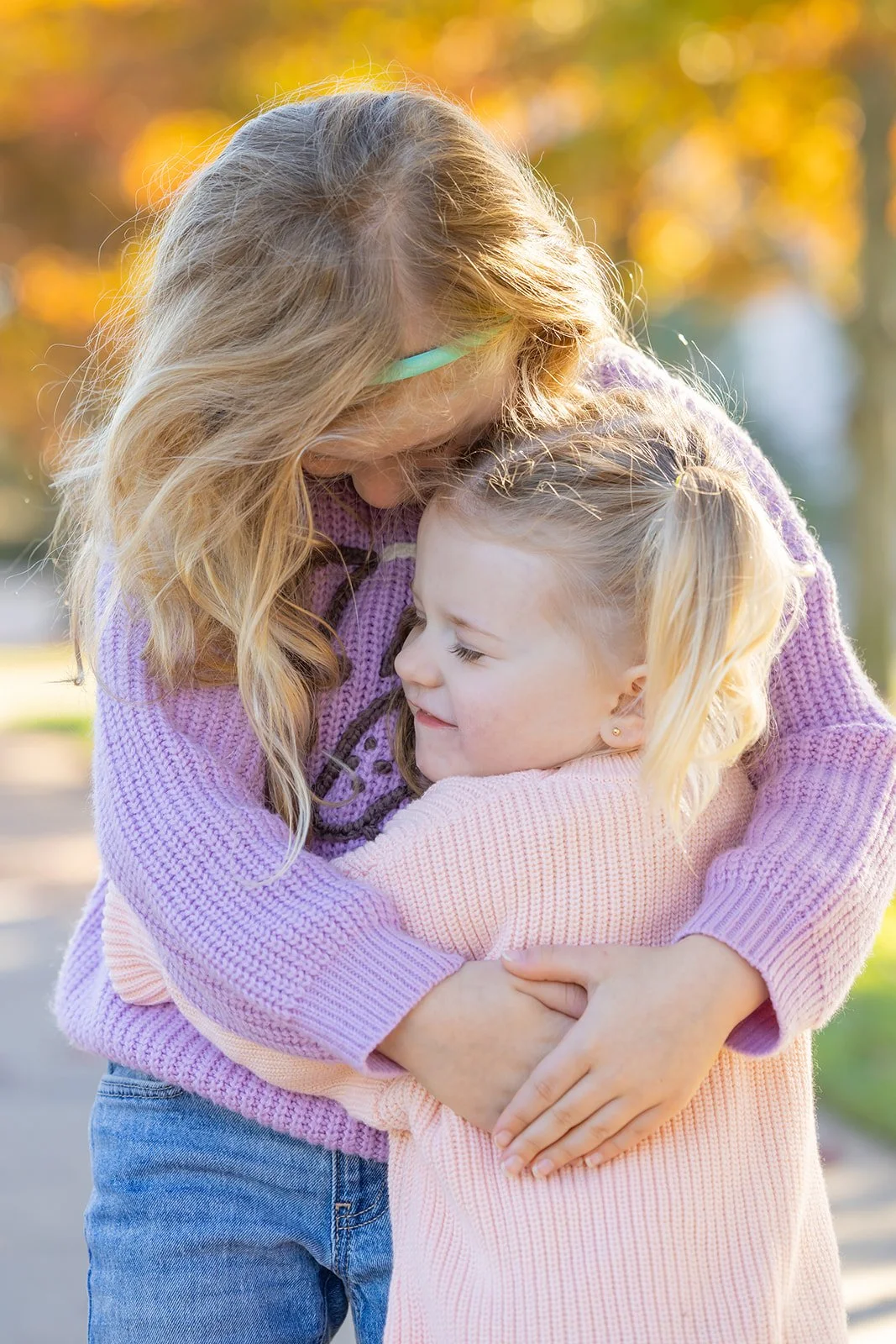 A woman with blonde hair wearing glasses hugging a young girl with blonde hair in a ponytail, both smiling, outdoors with autumn foliage in the background.
