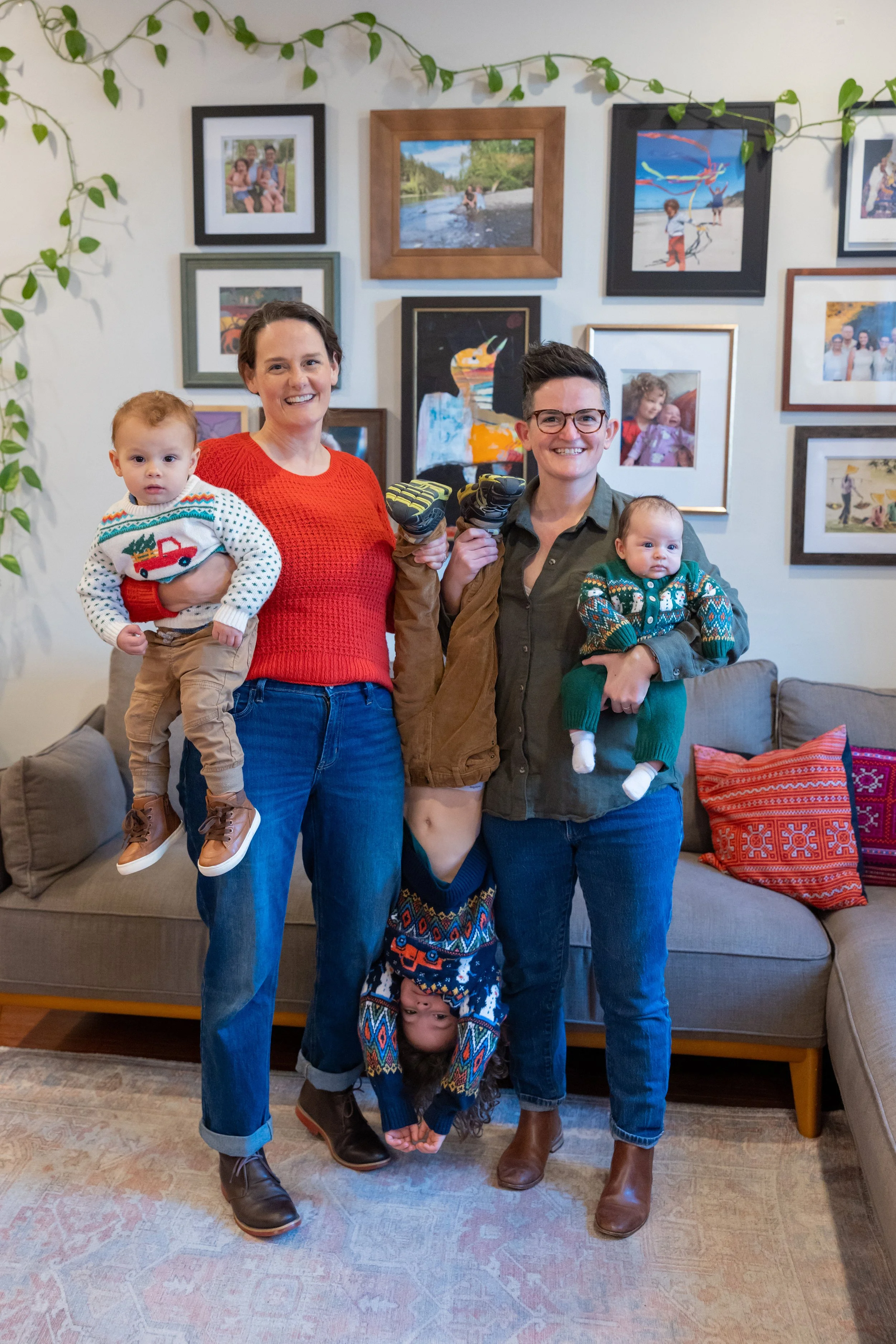 Two women and two children standing in a living room with a wall decorated with framed photos and artwork in the background. The women are smiling, holding small children, and making playful poses with their arms. One woman is wearing a red sweater, 