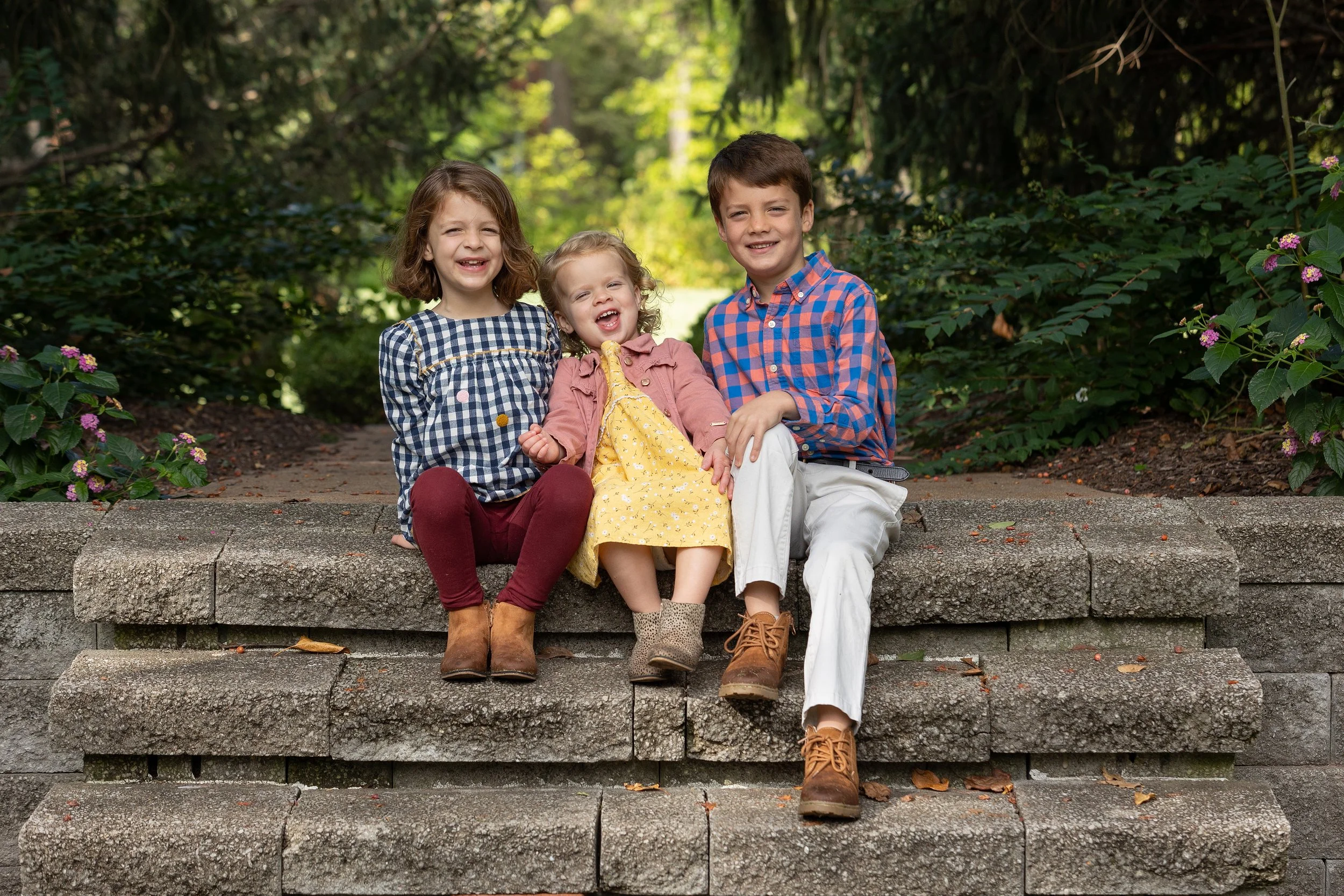 Three children sitting on stone stairs outdoors, smiling and posing for the camera, surrounded by greenery and flowers.