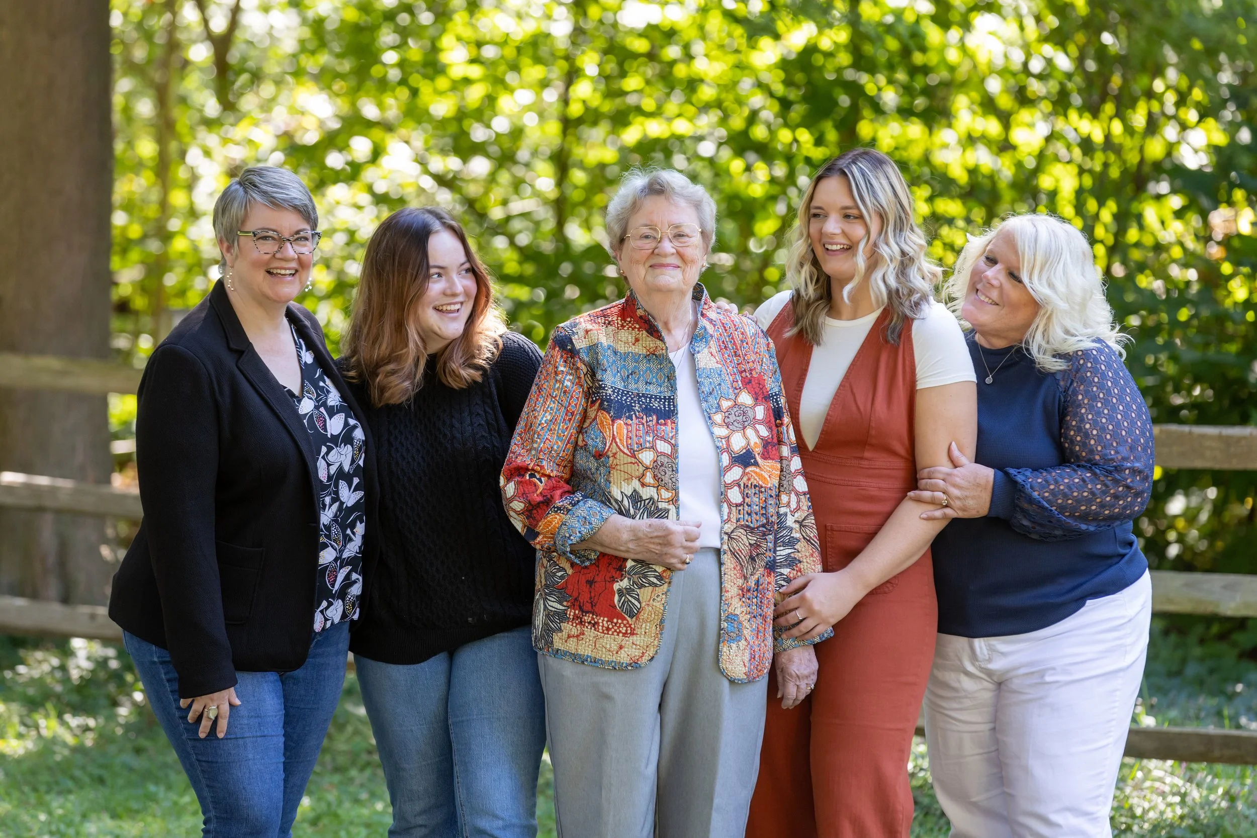 A group of five women standing outdoors in a park with green foliage in the background, smiling and posing for the camera.