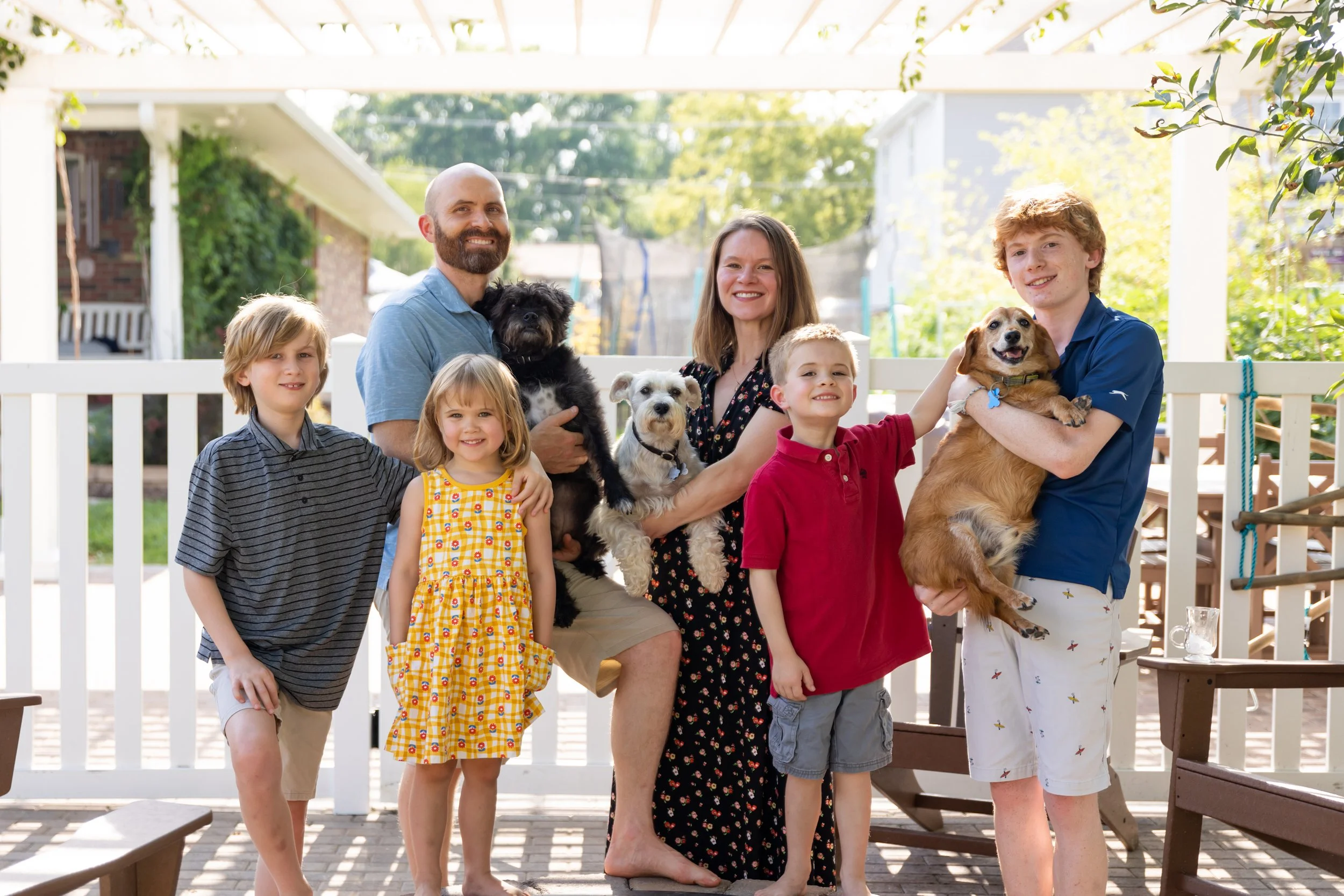 A family of seven with four children, two women, and a man, posing outdoors on a patio with their four dogs during daytime.