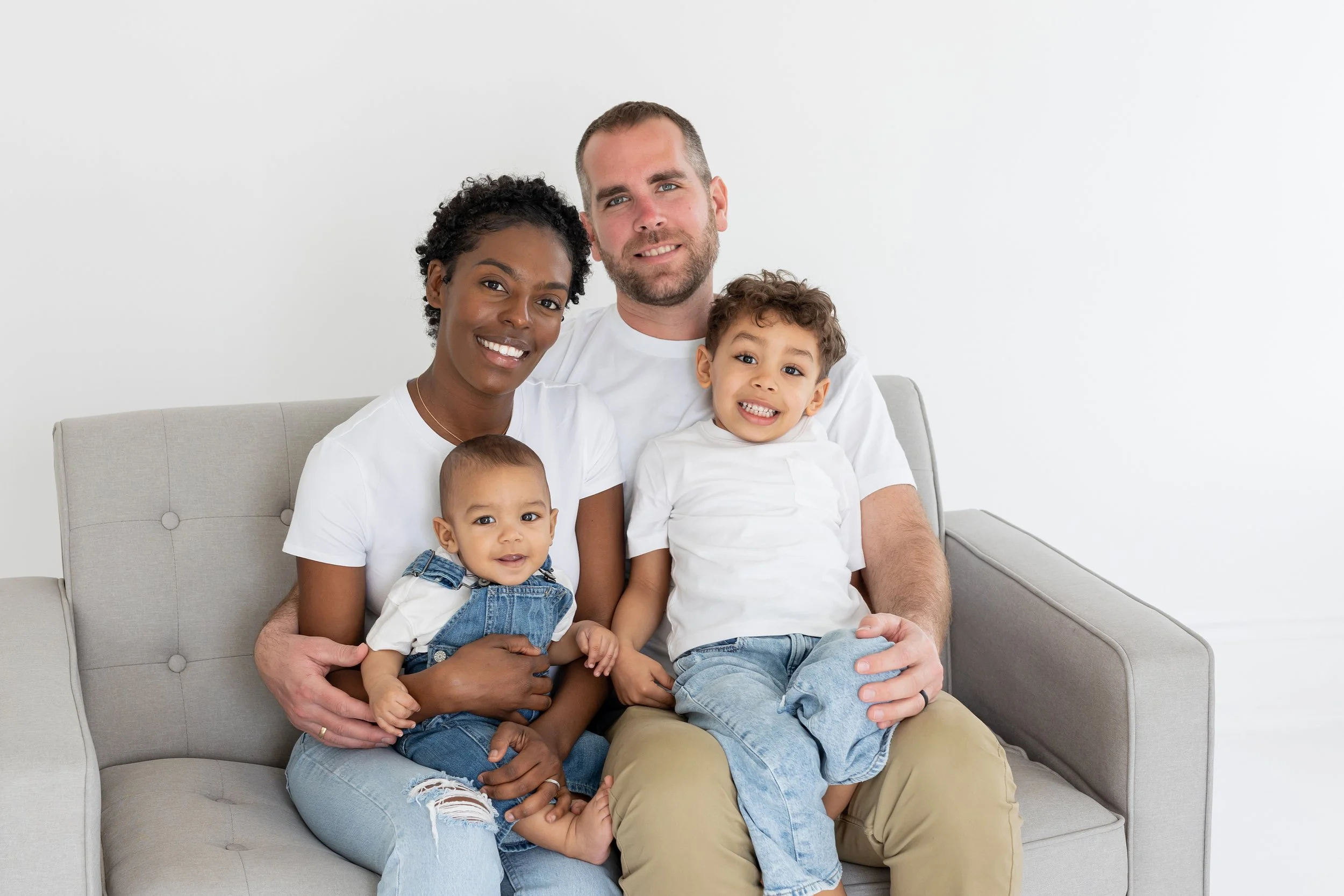 Family of five sitting on a light gray sofa in a bright room, smiling at the camera. The family includes a woman with dark curly hair, a man with short brown hair and beard, and three children—a baby, a young girl, and a young boy, all wearing casual