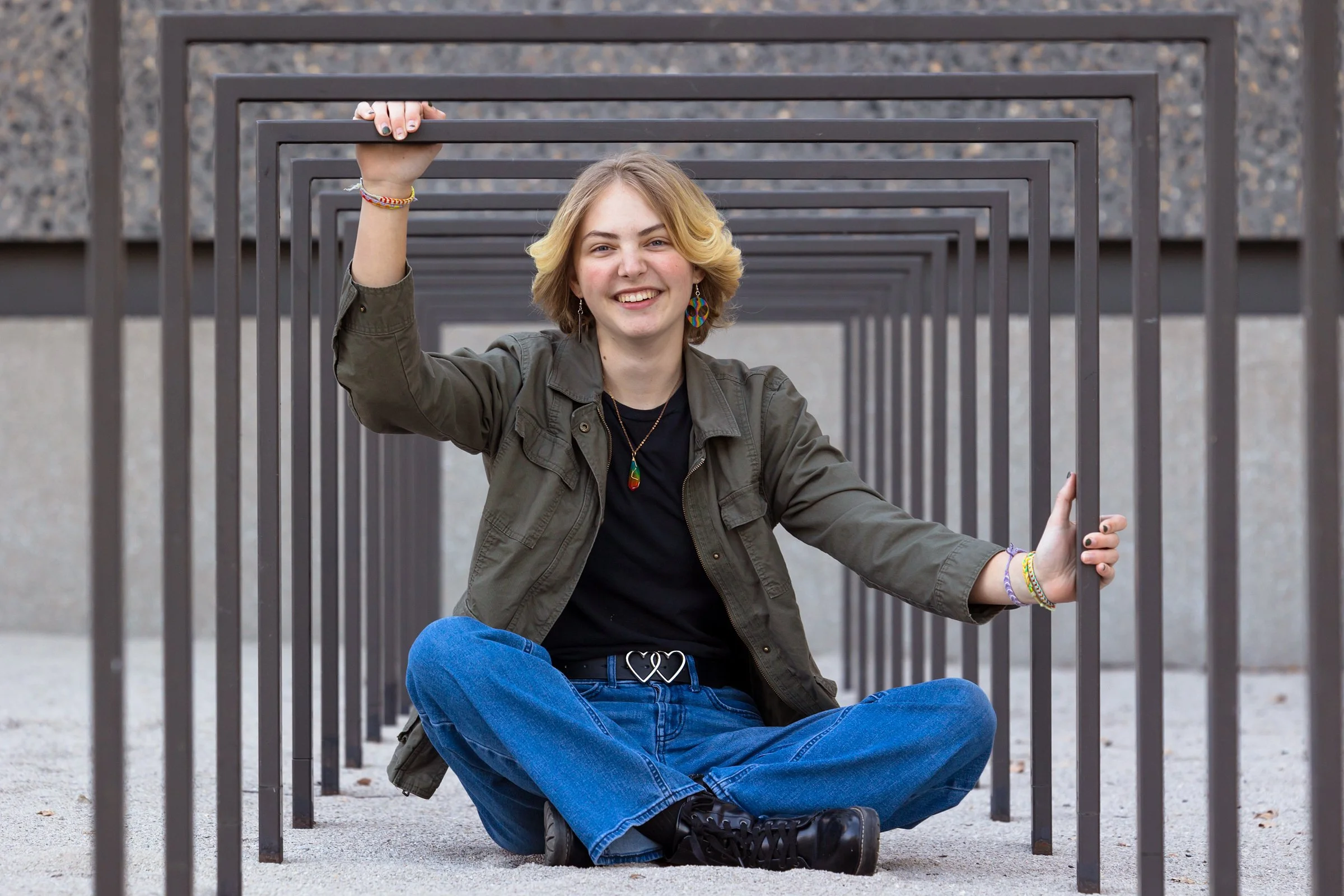 A young woman sitting on the ground, holding onto a metal frame structure, smiling at the camera. She has blonde hair, wears a dark green jacket, black shirt, blue jeans with a heart-shaped belt buckle, and black boots. She is also wearing colorful b