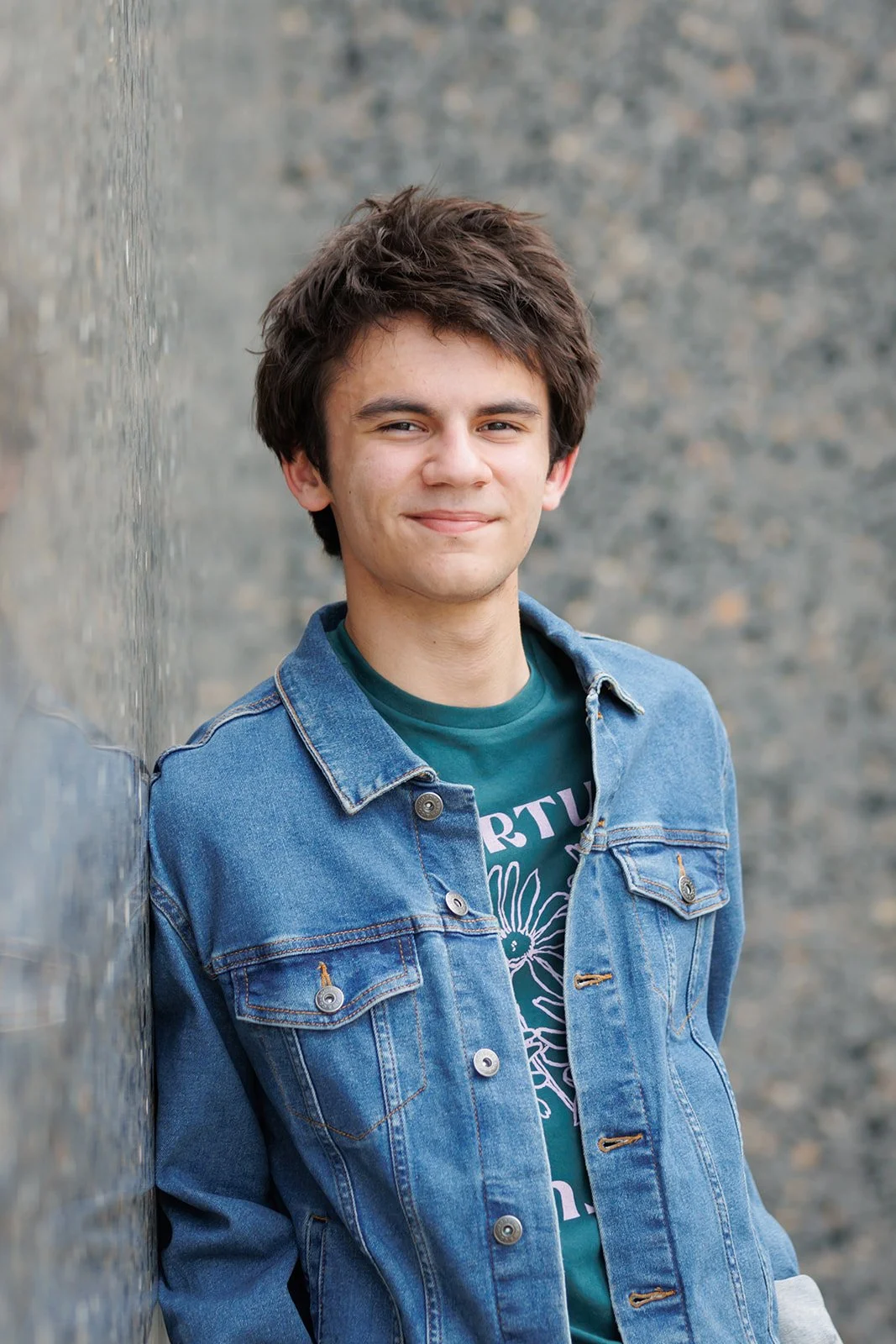 Young man with dark hair smiling, wearing a denim jacket over a graphic T-shirt, leaning against a textured gray wall.