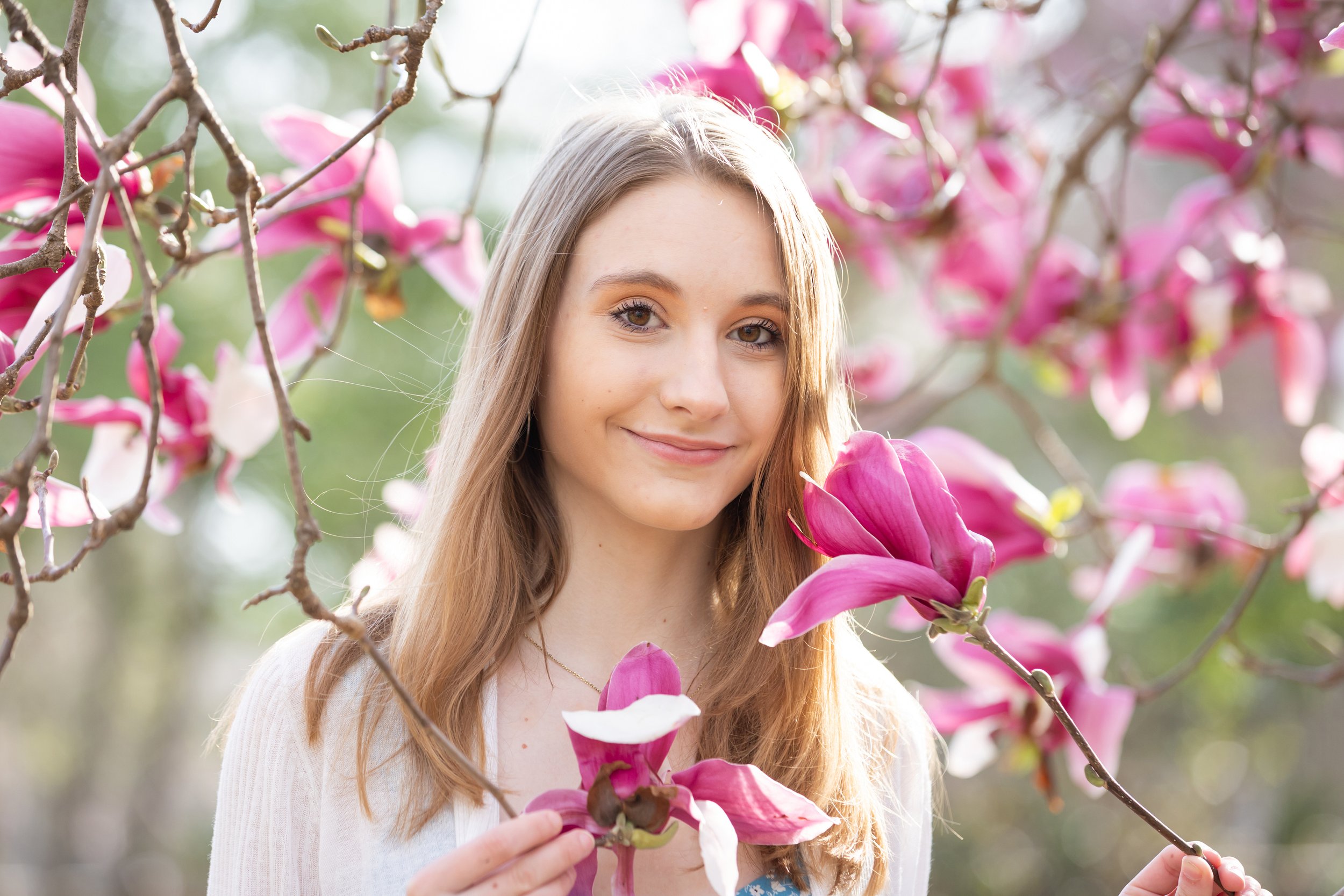 A young woman with long blonde hair smiling outdoors among pink flowers on tree branches, posing for her high school senior portraits