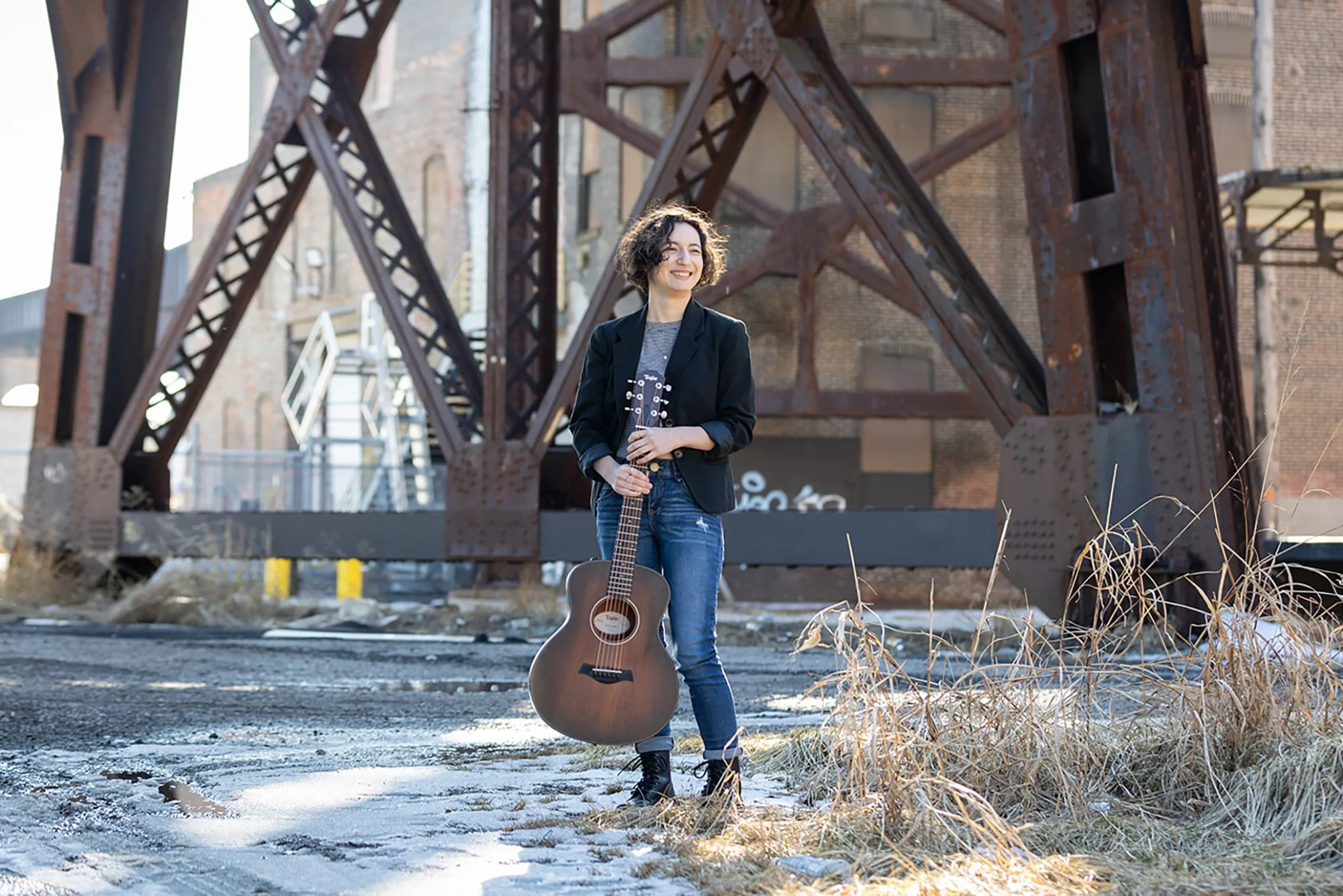 A woman standing outdoors on a cold day holding an acoustic guitar, smiling, with an old rusty iron bridge and brick building in the background.