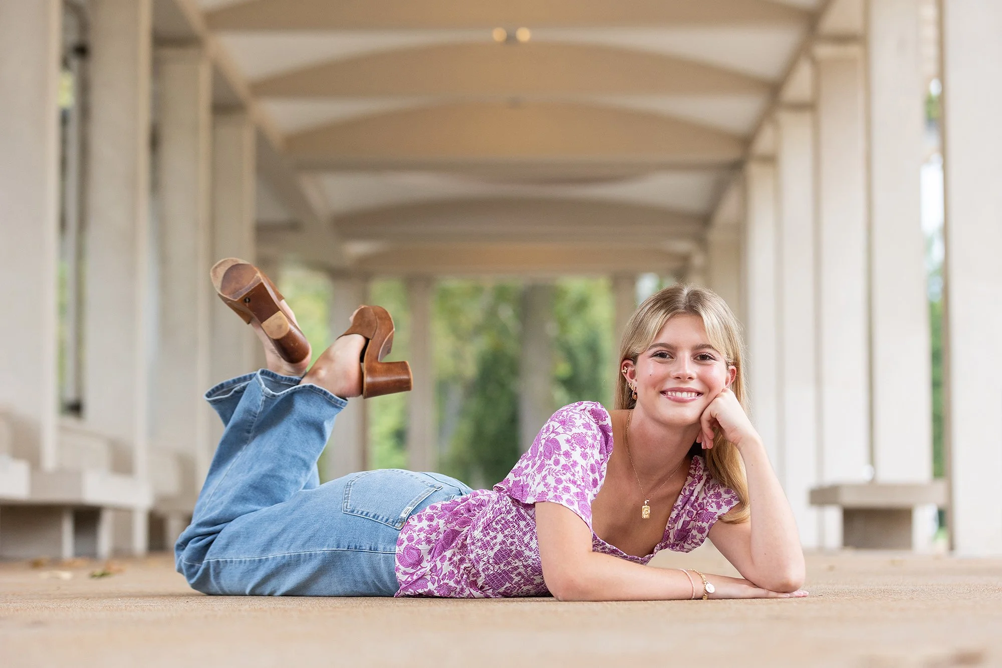 Young woman with blonde hair lying on her stomach on a carpeted walkway under a pavilion, smiling and resting her chin on her hand, wearing a pink patterned top, blue jeans, and brown sandals.