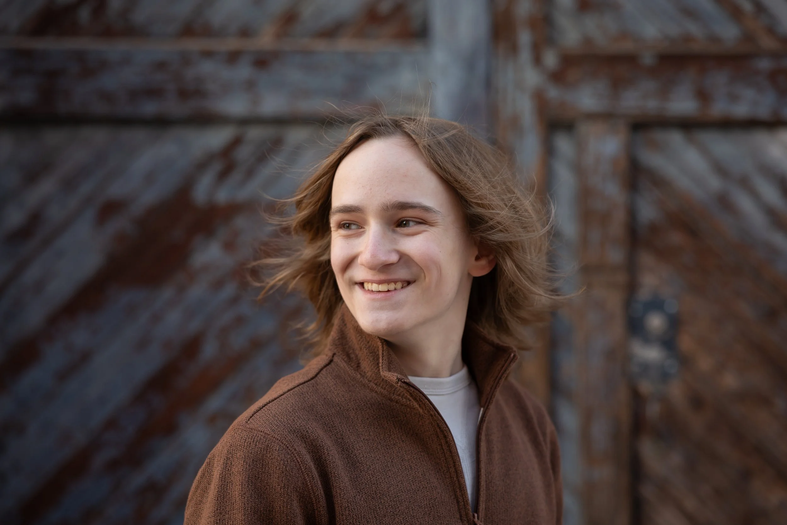 Young man with shoulder-length hair smiling outdoors, wearing a brown jacket and a white shirt, standing in front of a weathered wooden fence.