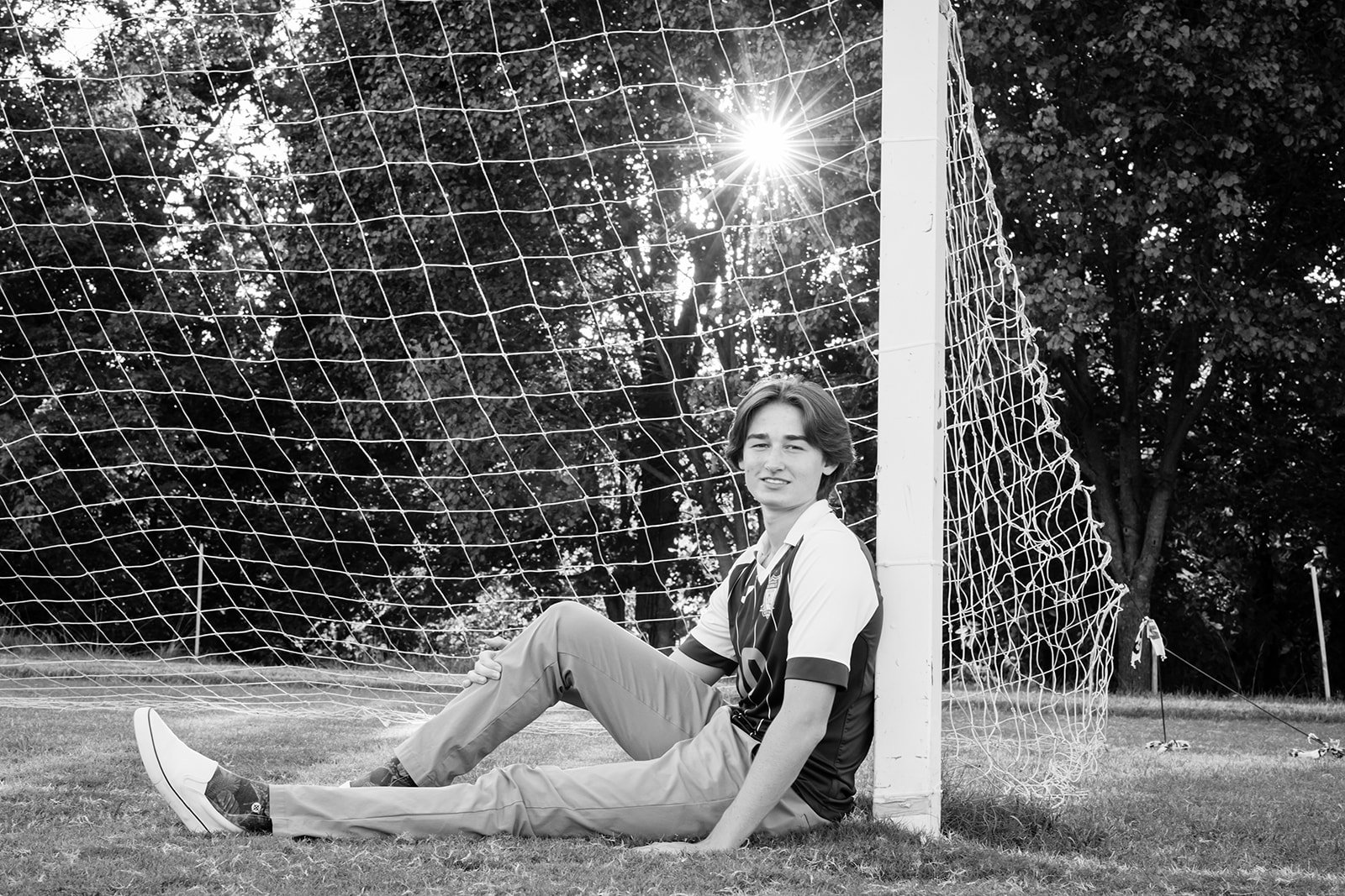 A young male soccer player sitting on the grass next to a goalpost on a soccer field, with sunlight shining through the trees in the background.
