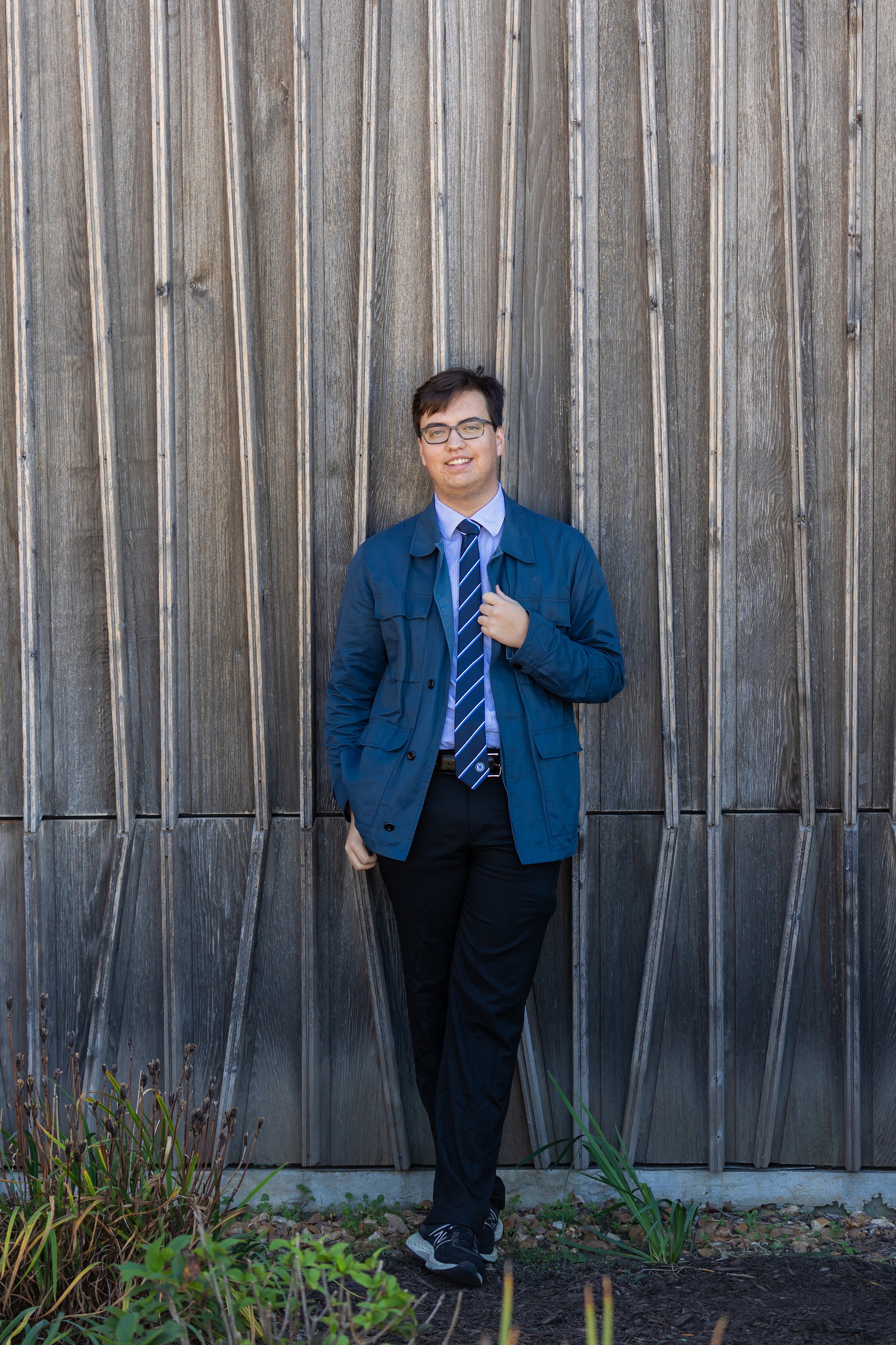 A young man with glasses, dressed in a blue jacket, dress shirt, and striped tie, standing against a wooden fence outdoors.