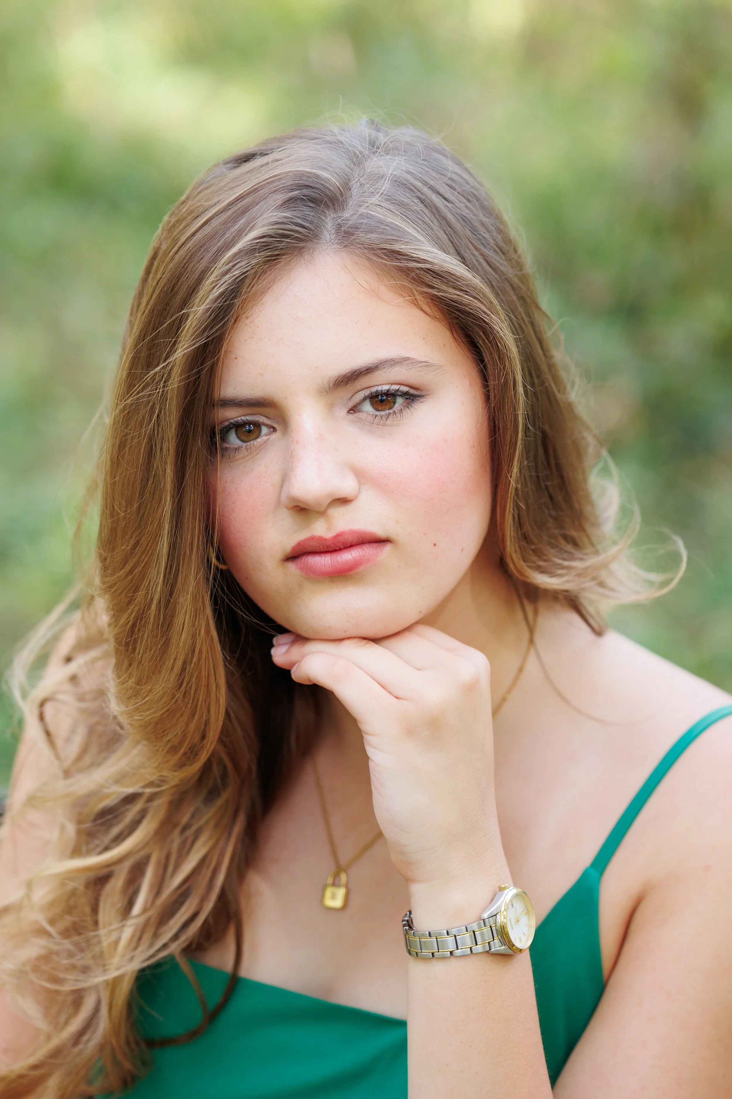 Close-up of a young woman with long, wavy brown hair, fair skin, and brown eyes, wearing a green tank top and a silver watch, resting her chin on her hand outdoors.