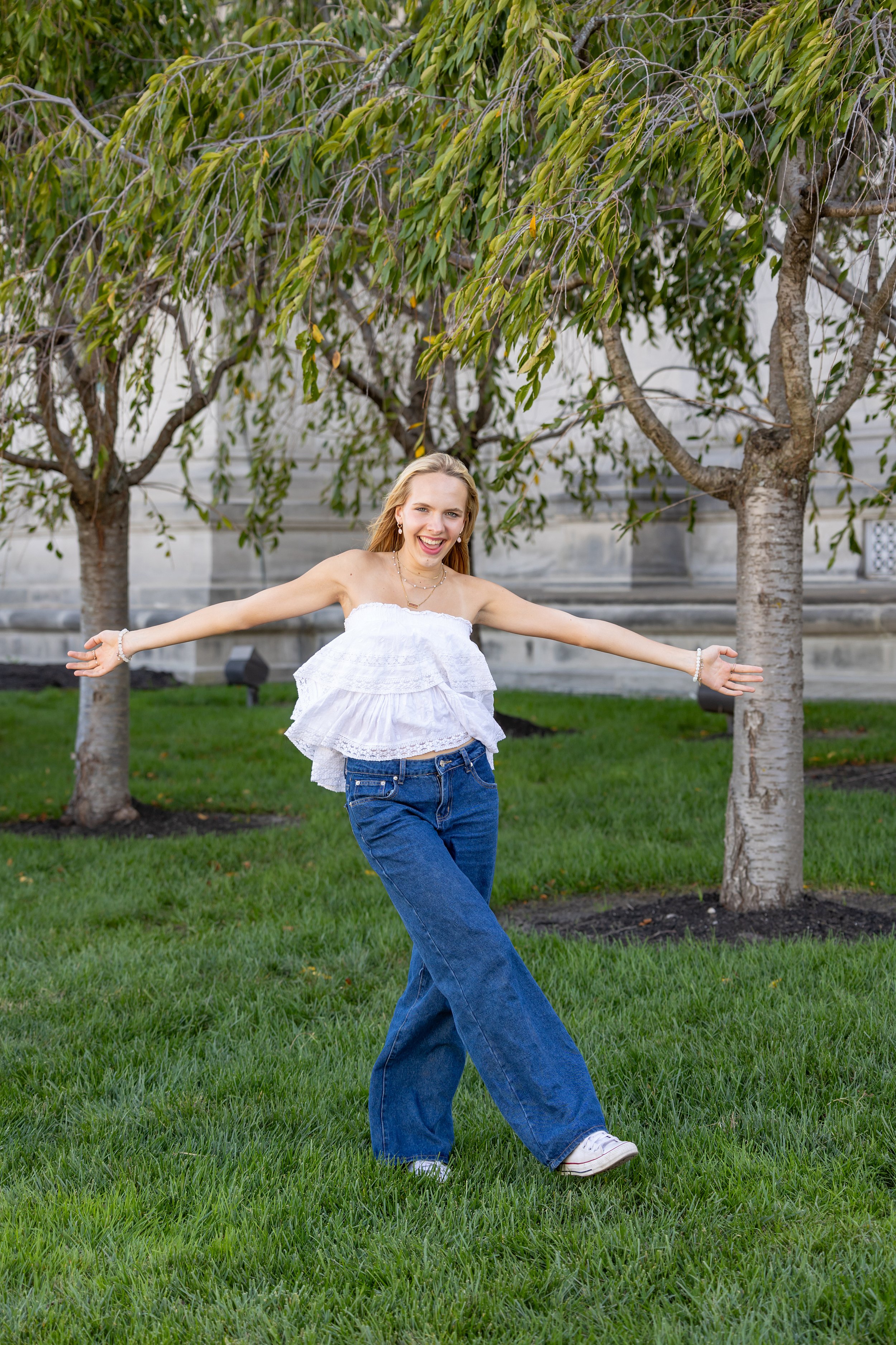 A young woman with long blonde hair smiling and standing with arms outstretched on a grassy lawn, wearing a white sleeveless ruffled top, blue jeans, and white sneakers, with trees and a building in the background.